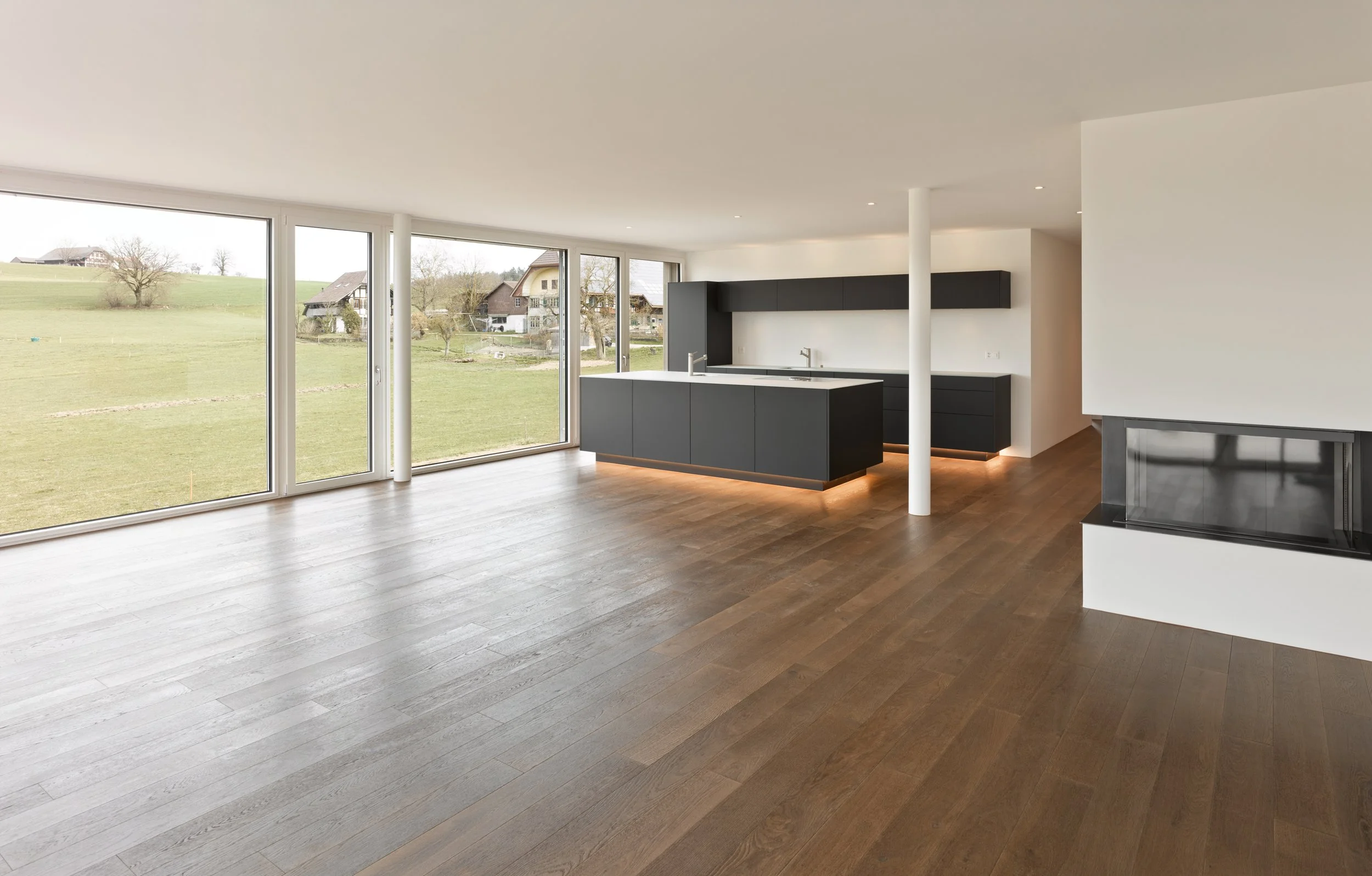 Ein modernes, leeres Wohnzimmer mit großen Fenstern, die eine grüne Landschaft mit Häusern im Hintergrund zeigen, und einer schwarzen Kücheninsel in der Mitte.