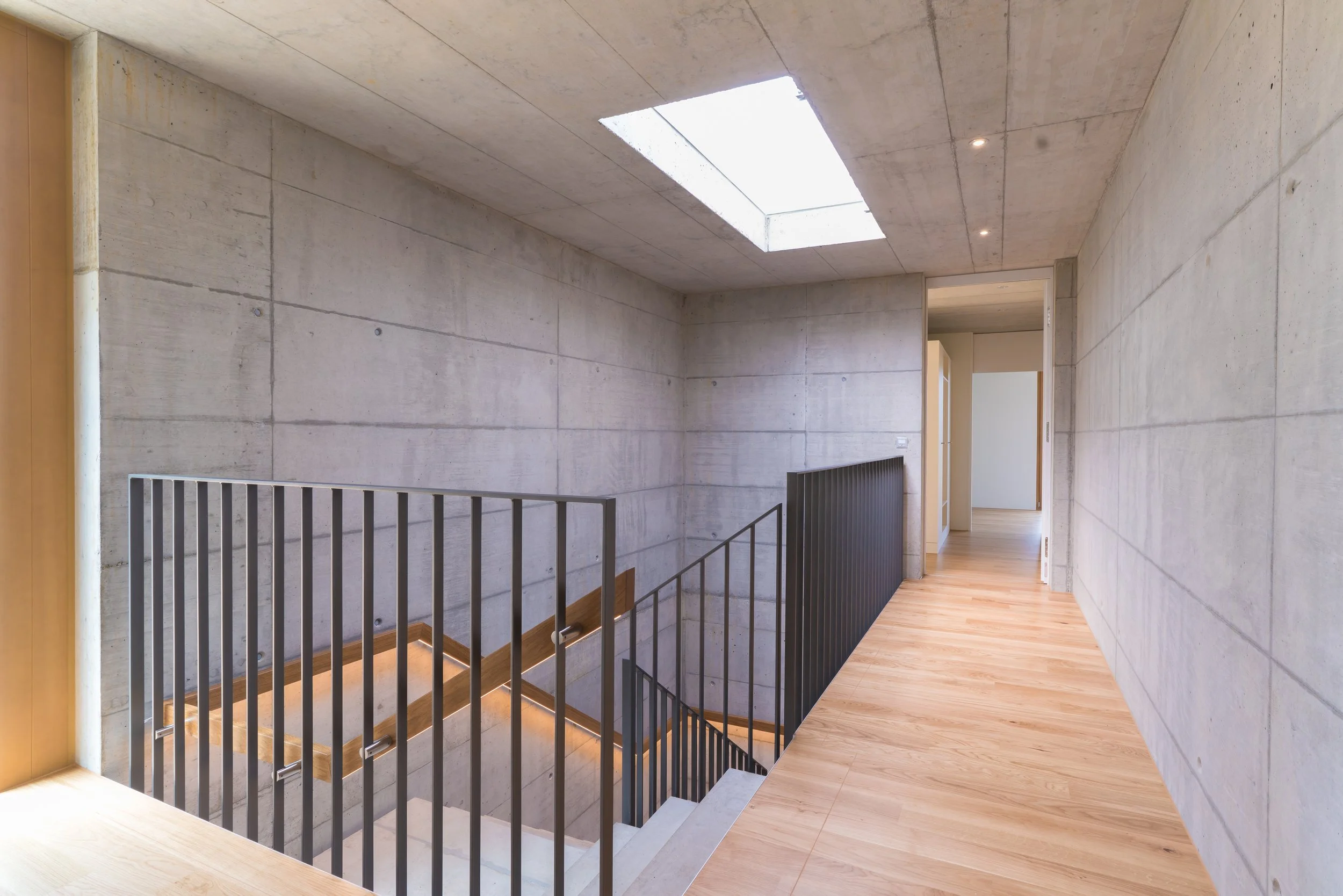 Innenansicht eines modernen Treppenhauses mit Betonwänden, Holzboden und einem großen Oberlicht in der Decke.