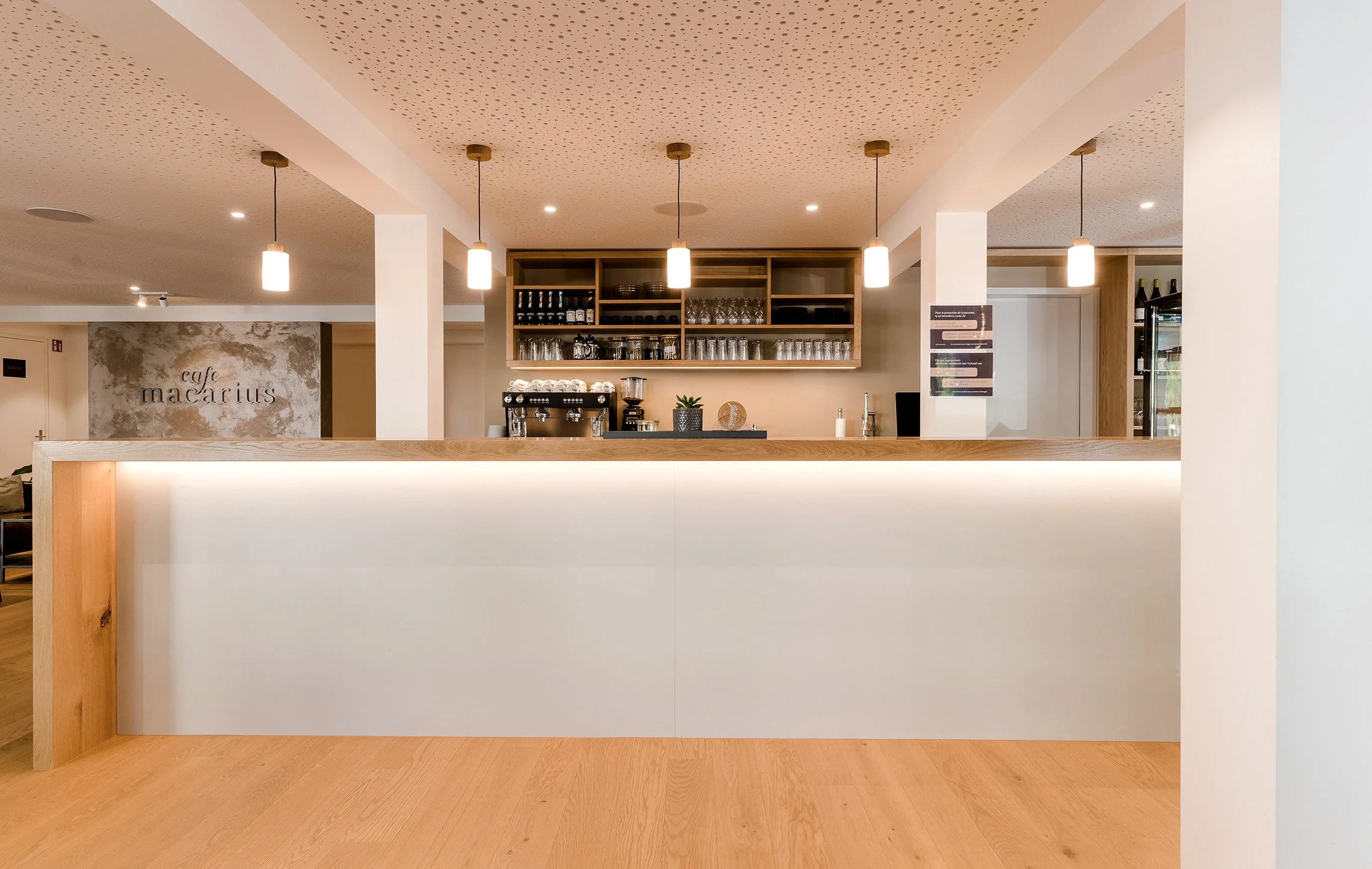Innenansicht einer modernen, minimalistisch eingerichteten Café-Bar mit hellem Holz und weißen Wänden, beleuchtet von vier hängenden Lampen, mit einer Bar und einem Regal, das Gläser und Flaschen enthält.