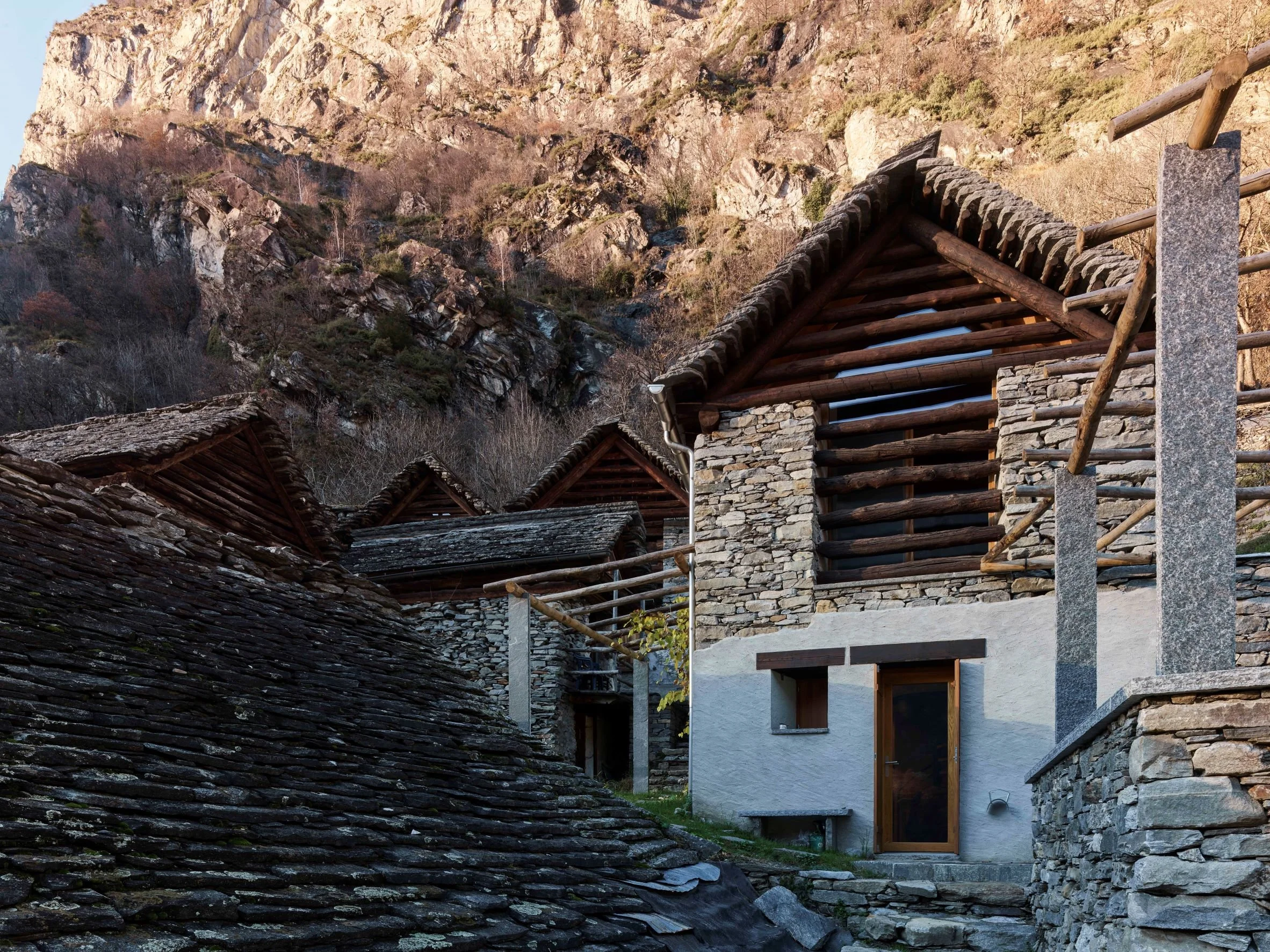 Traditionelle Bergbauernhäuser aus Stein und Holz in einer alpinen Landschaft mit Felsen und Bäumen im Hintergrund.