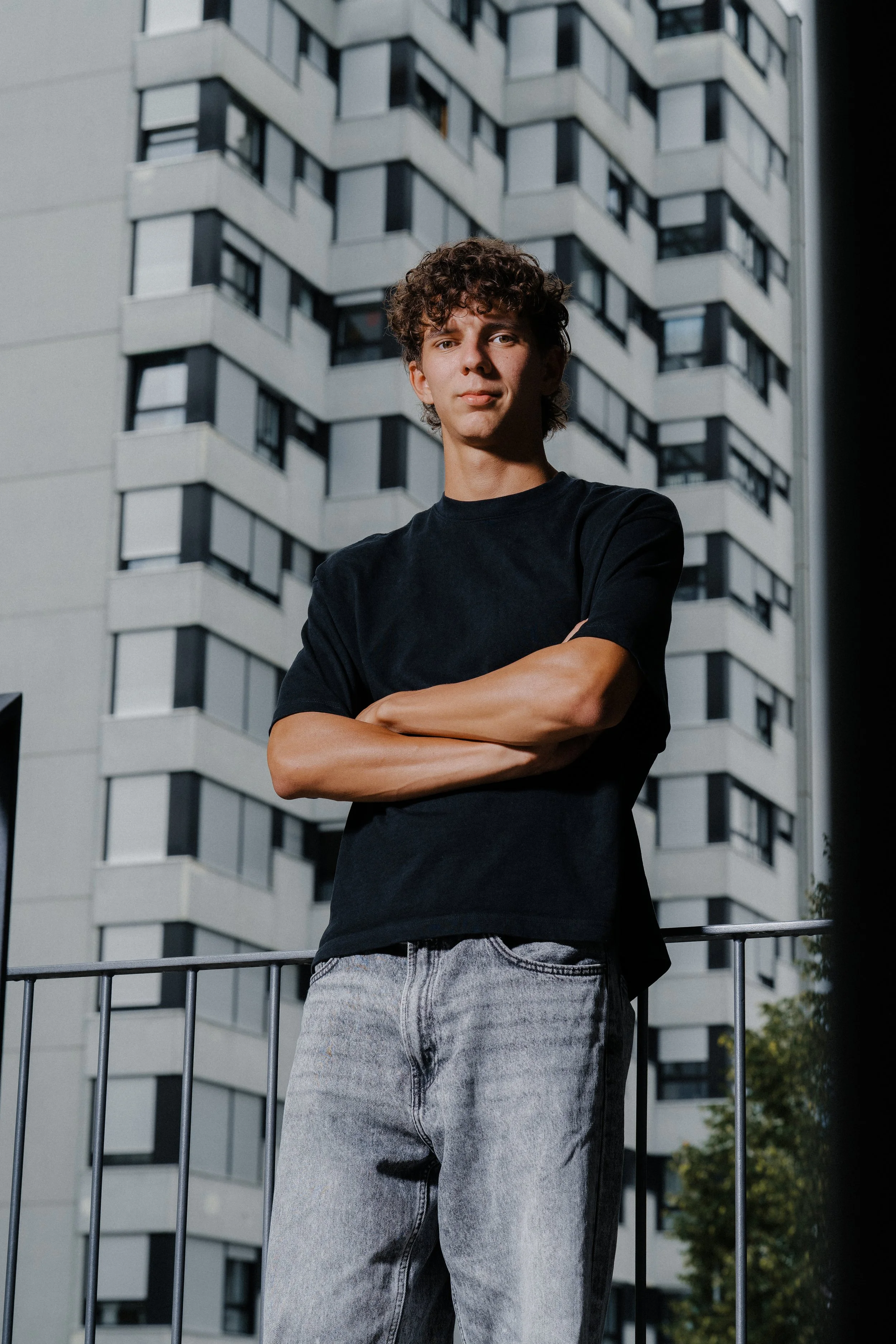 Junge Person mit verschränkten Armen in urbaner Umgebung an Hochhaus-Kulisse bei Tag.