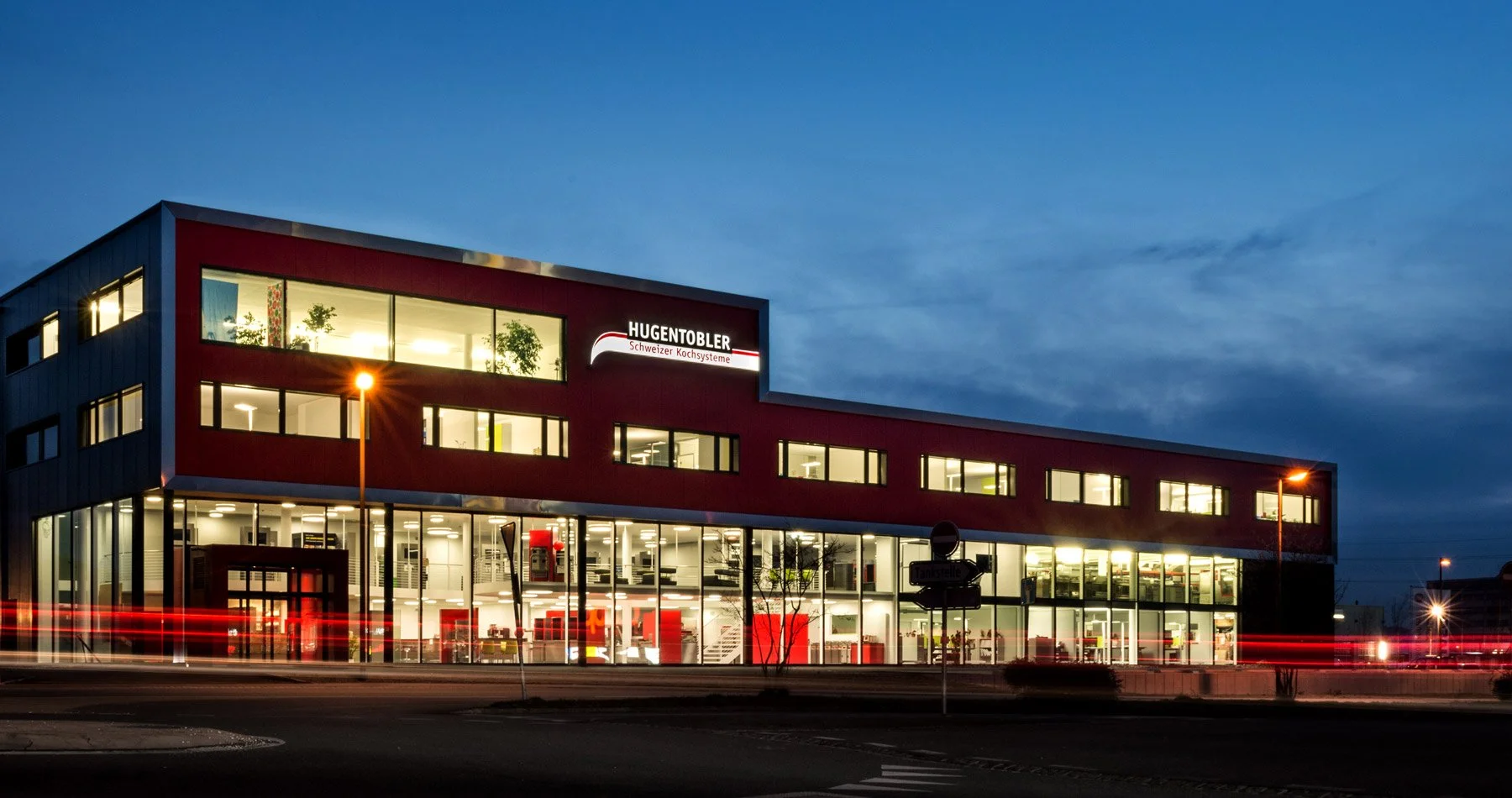 Bürogebäude bei Nacht mit beleuchteten Fenstern und einem beleuchteten Firmenlogo 'HUGENTOBLER Schweizer Kochsysteme' an der Fassade.