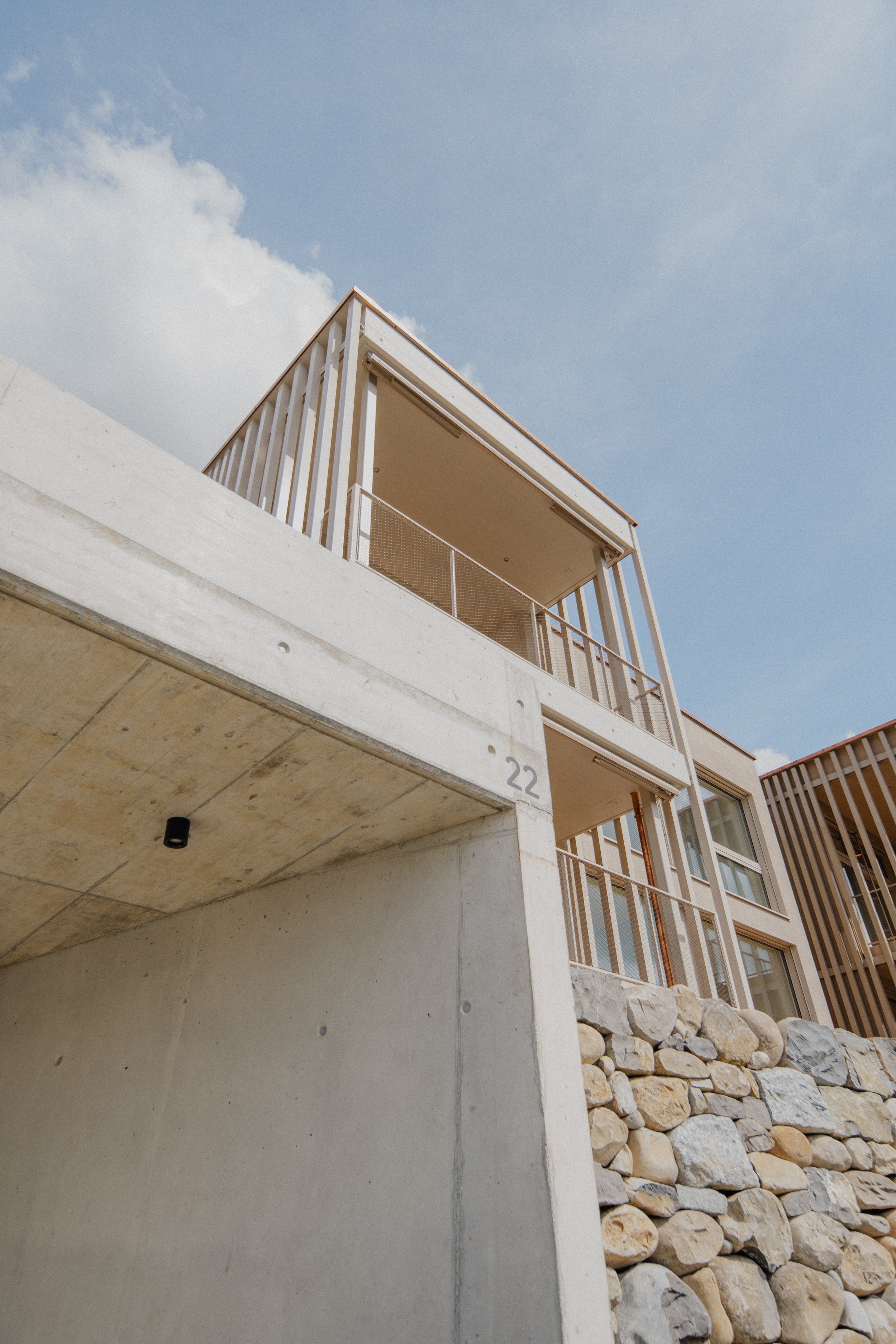 Aufnahme eines modernen Gebäudeteils mit Balkon, gelben Steinen, Beton und Holz, unter einem blauen Himmel mit Wolken.