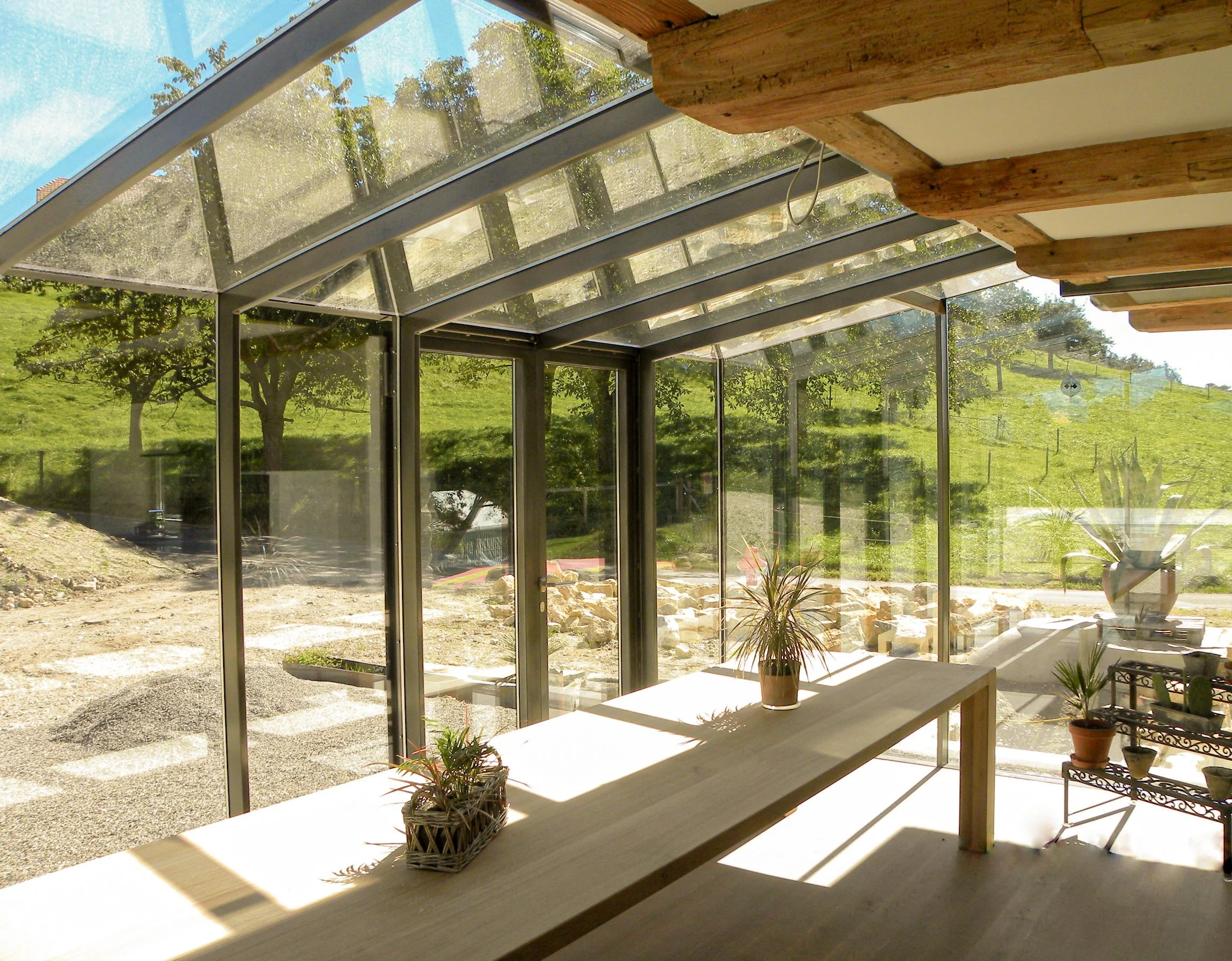 Innenansicht eines Hauses mit großen Glasfenstern, durch die man einen grünen Garten und Bäume sieht. Im Vordergrund steht eine Holzplatte mit Topfpflanzen.