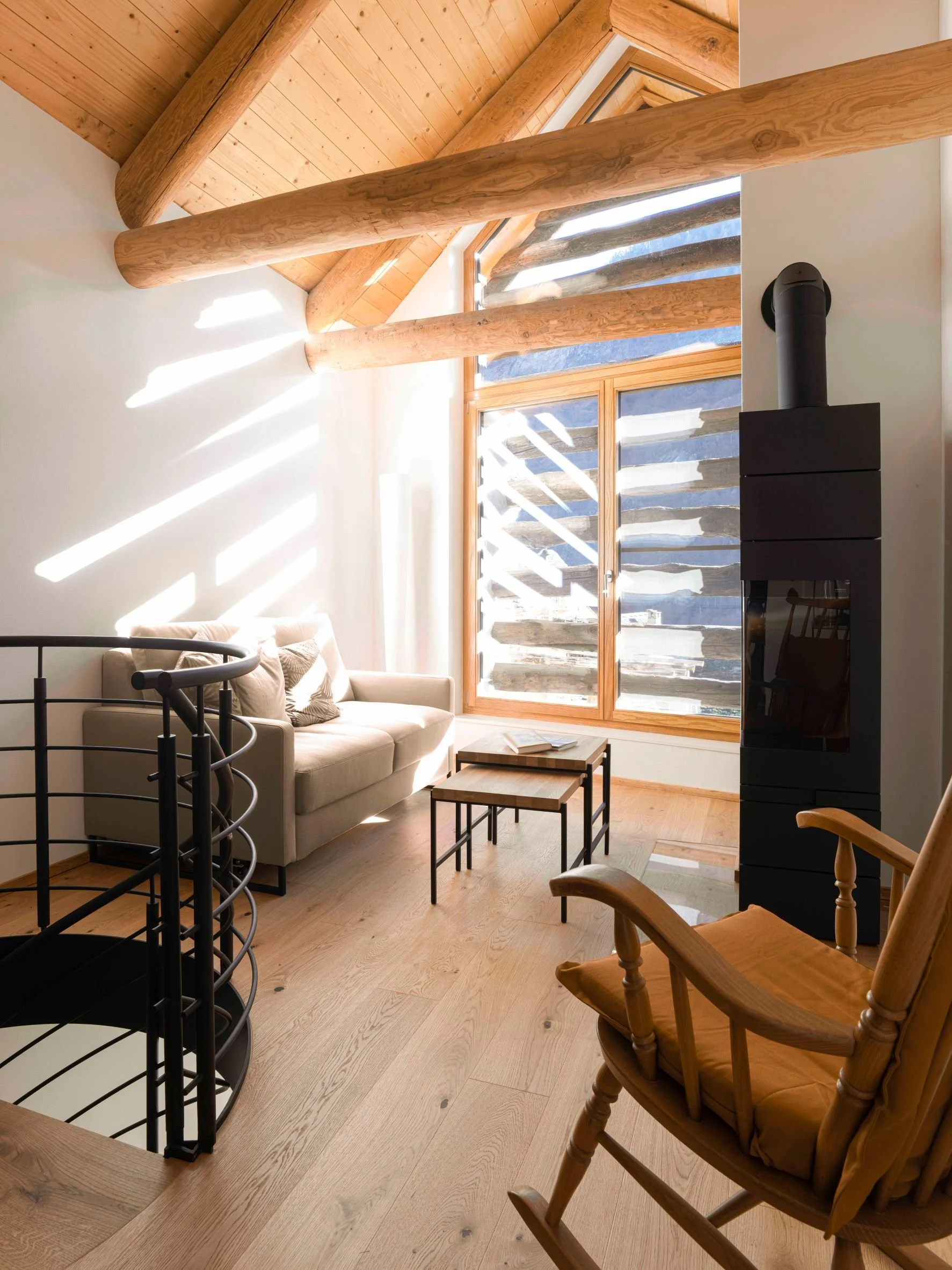 Gemütliches Wohnzimmer mit Holzdecke und großen Fenstern, Couchecke, Holzboden, Kamin, Holzstuhl, und moderner schwarzen Kaminofen.