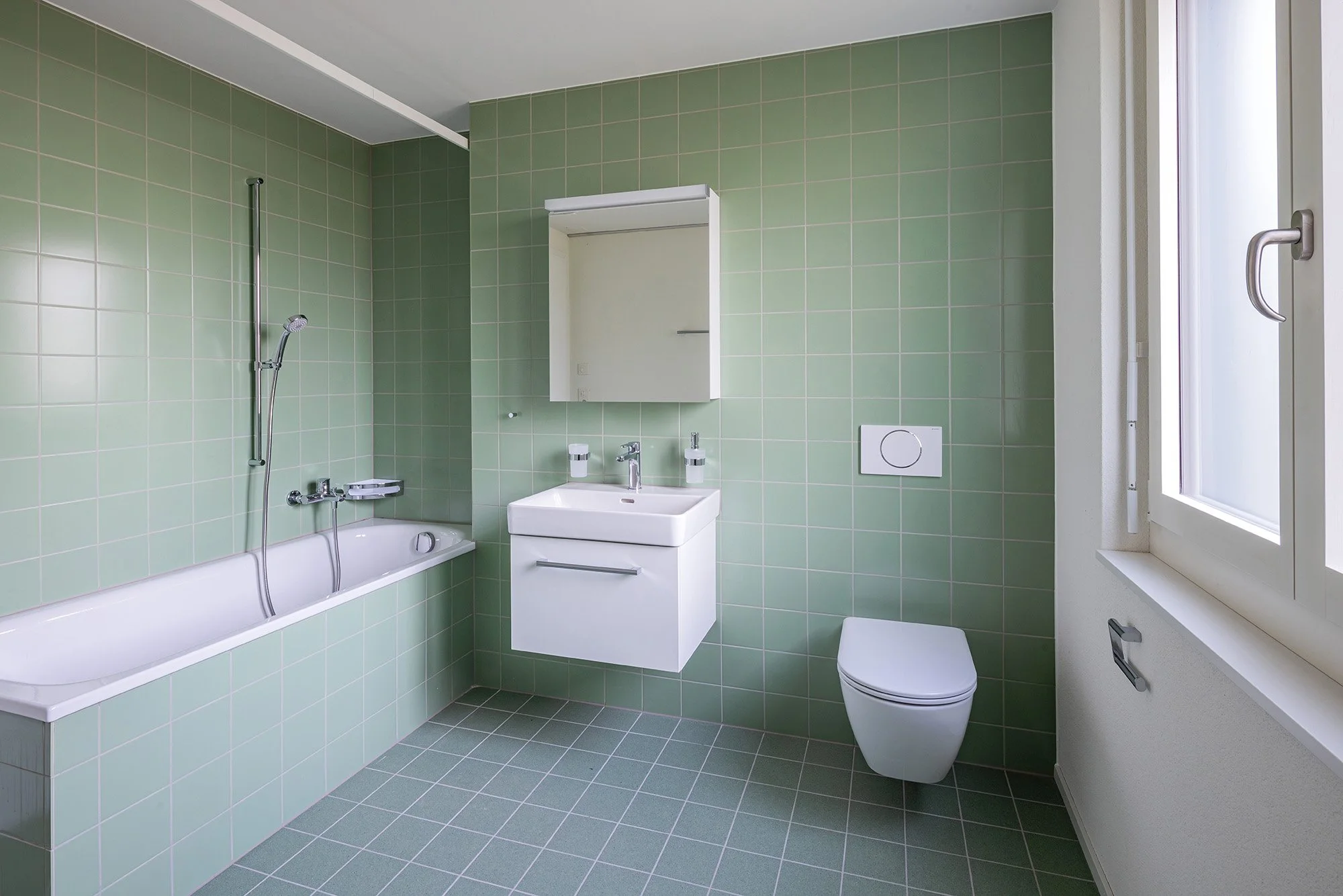 Ein modernes Badezimmer mit grünen Wandfliesen, einer Badewanne, einem Waschbecken mit Spiegel und einer Toilette.