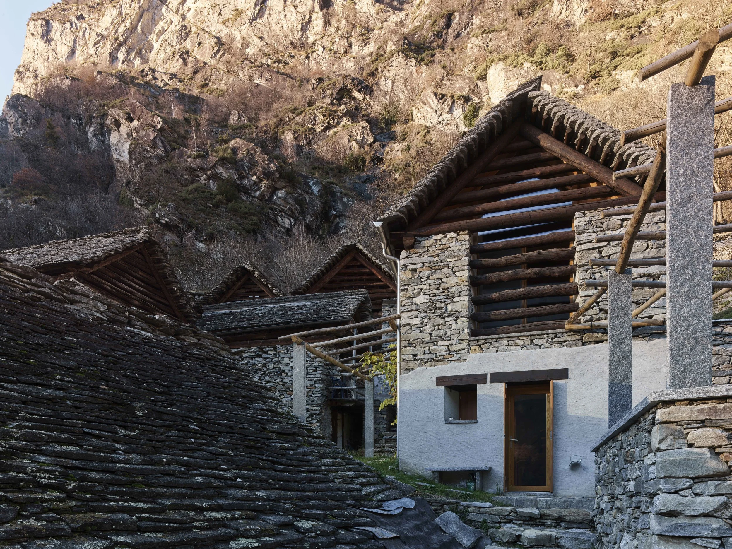 Traditionelle Steinhäuser mit HolzDach in einer Bergregion, umgeben von Felsen und Bäumen im Winter.
