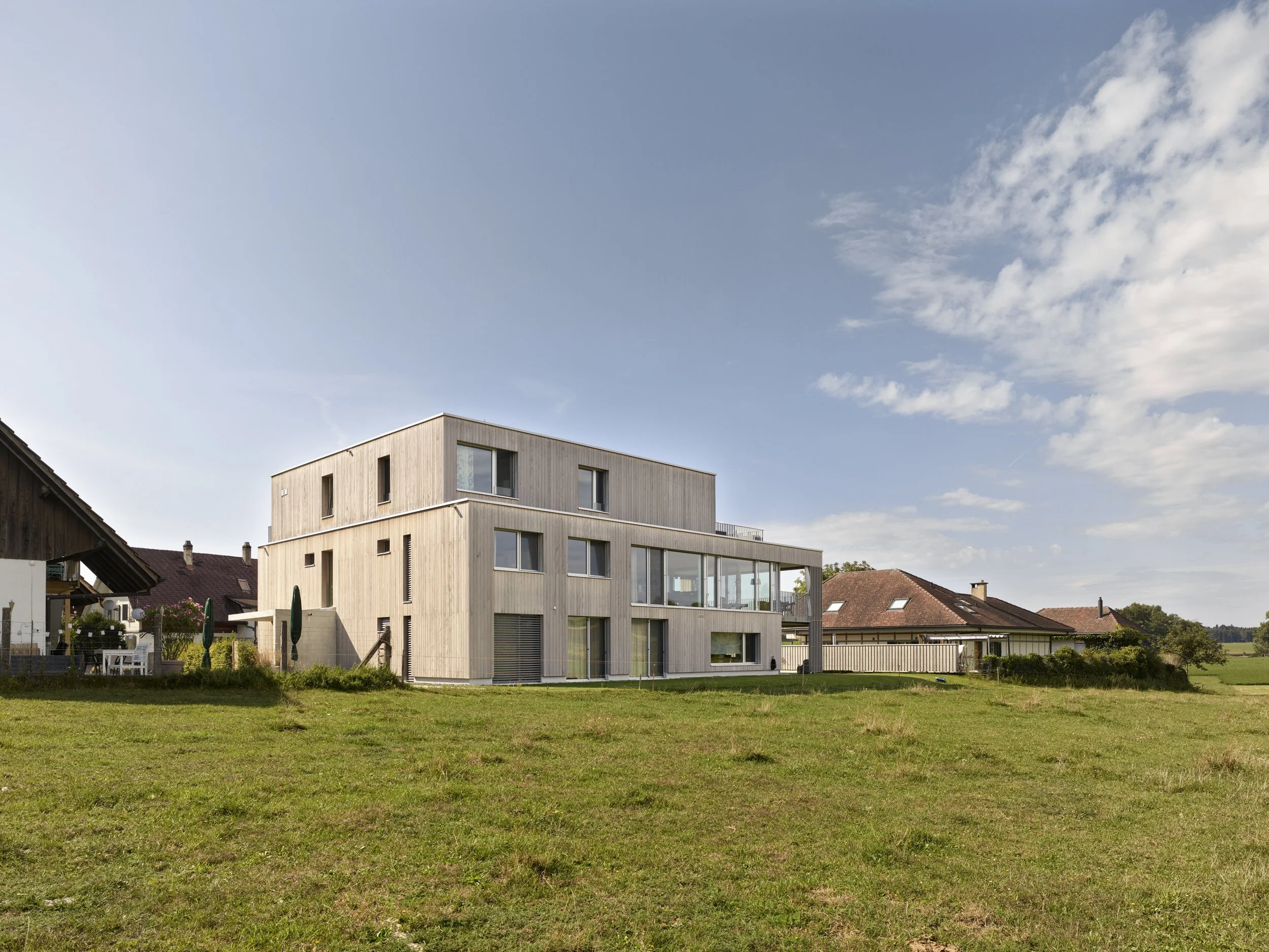 Modernes mehrstöckiges Wohnhaus mit großen Fenstern und Holzwandfassade, umgeben von einem grünen Rasen, bei blauem Himmel mit Wolken.