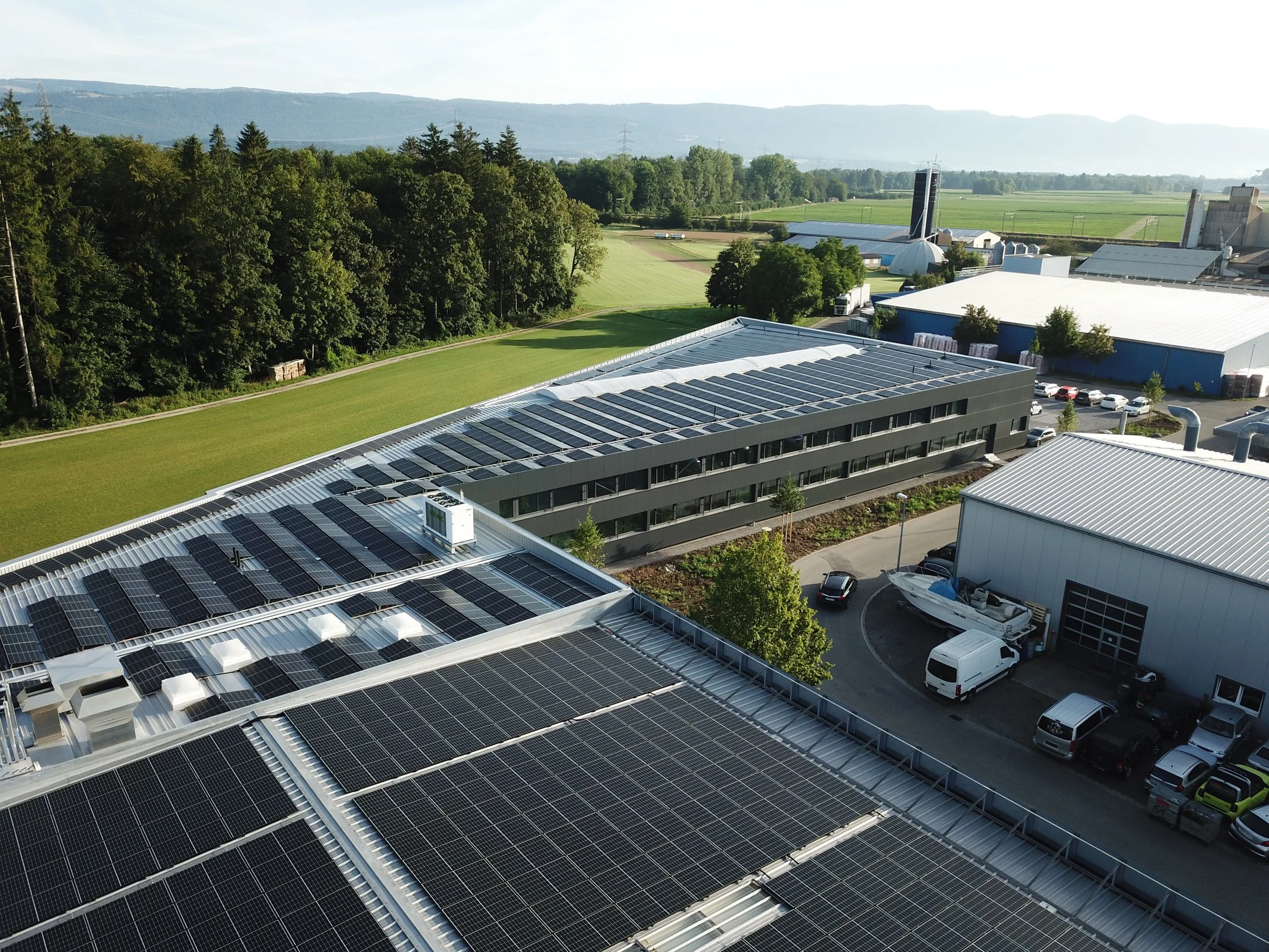 Modernes Gebäude mit Solaranlagen auf dem Dach, umgeben von Bäumen und landwirtschaftlichen Flächen im Hintergrund.