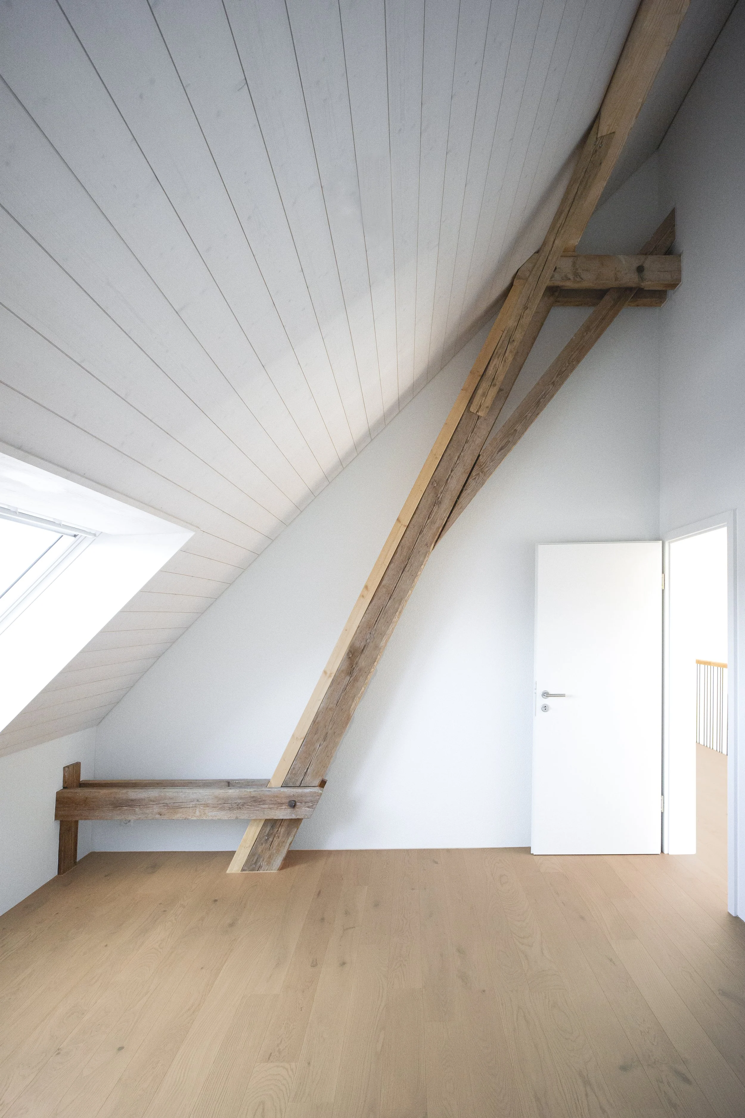 Innenraum mit weiß gestrichener Wand, gekreuztem Holzbalken und einer weißen Tür, Fenster im Dach, Holzboden.