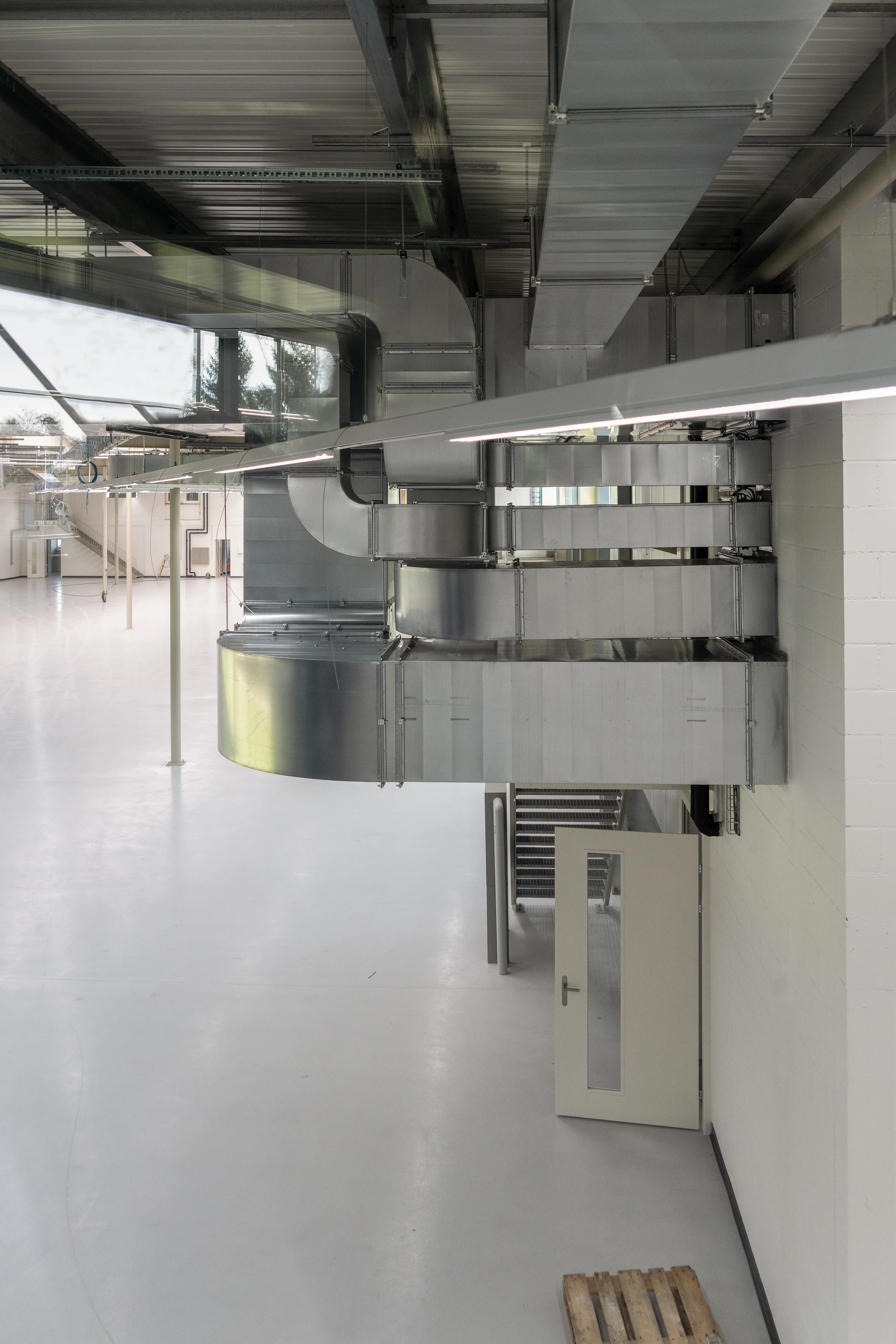 Innenraum einer Industriehalle mit großen metallischen Lüftungsrohren, weißen Wänden, einem Glastor mit Blick nach draußen und einer weißen Verteilerbox auf dem Boden.