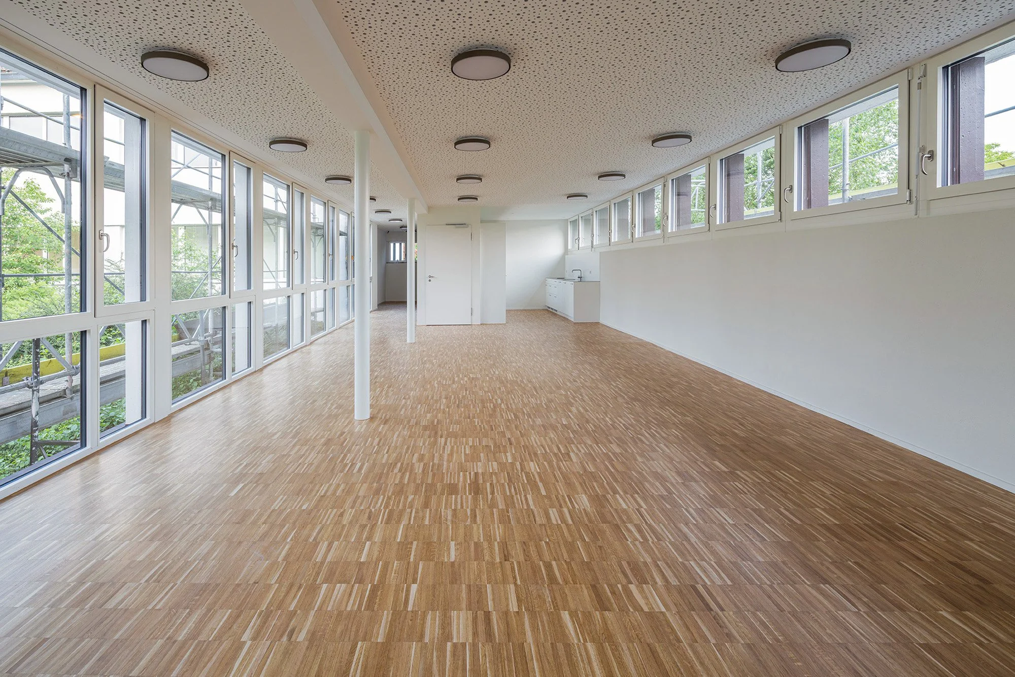 Leeres Zimmer mit großen Fenstern, Holzboden, weißen Wänden und Deckenleuchten.