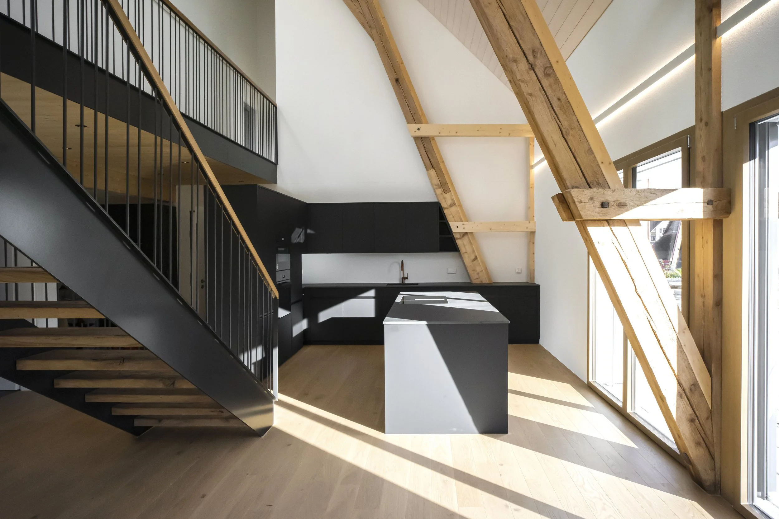 Innenaufnahme eines modernen Raumes mit sichtbaren Holz-Dachstrukturen, großen Fenstern, schwarzer Treppe und schwarzem Küchenbereich.
