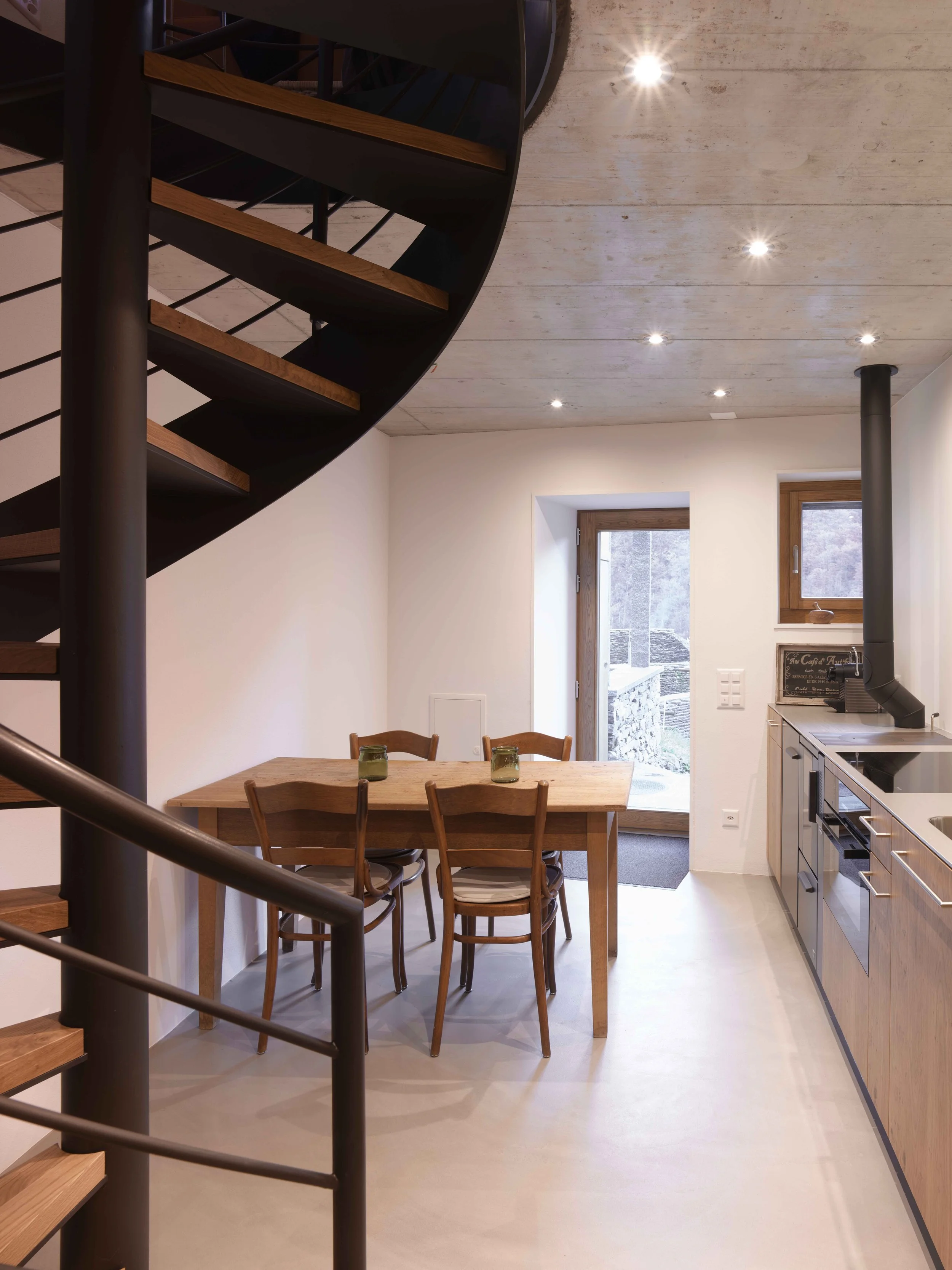 Innenansicht einer modernen Wohnung mit Holz-Esstisch, vier Stühlen, offener Küche mit Herd und Fenster nach draußen, beleuchtet mit Deckenleuchten und natürlichem Licht.