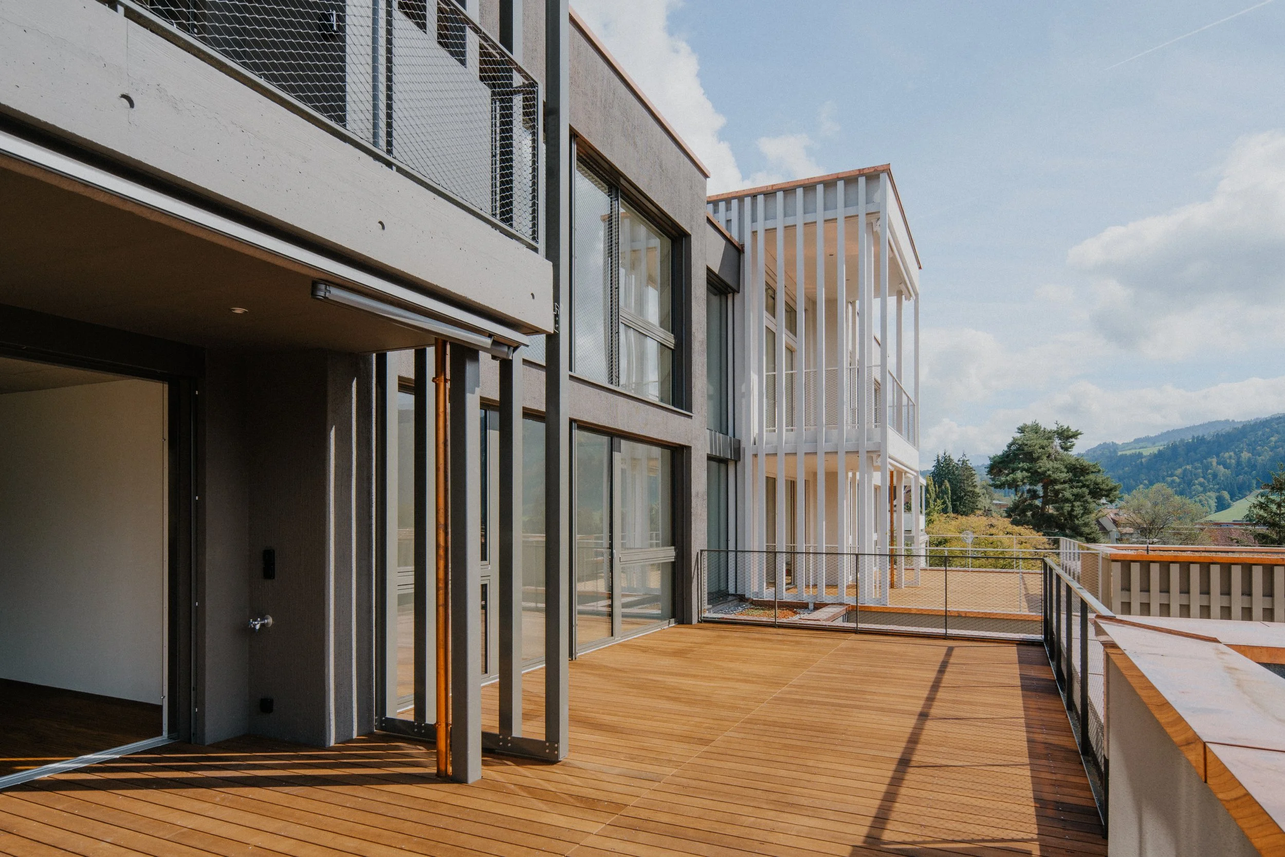 Balkon eines modernen Mehrfamilienhauses mit Holzdecks, Glas- und Metallgeländer sowie weiße vertikale Lamellen im Hintergrund, Blick auf eine Landschaft mit Bäumen und Hügeln bei Himmel mit Wolken.