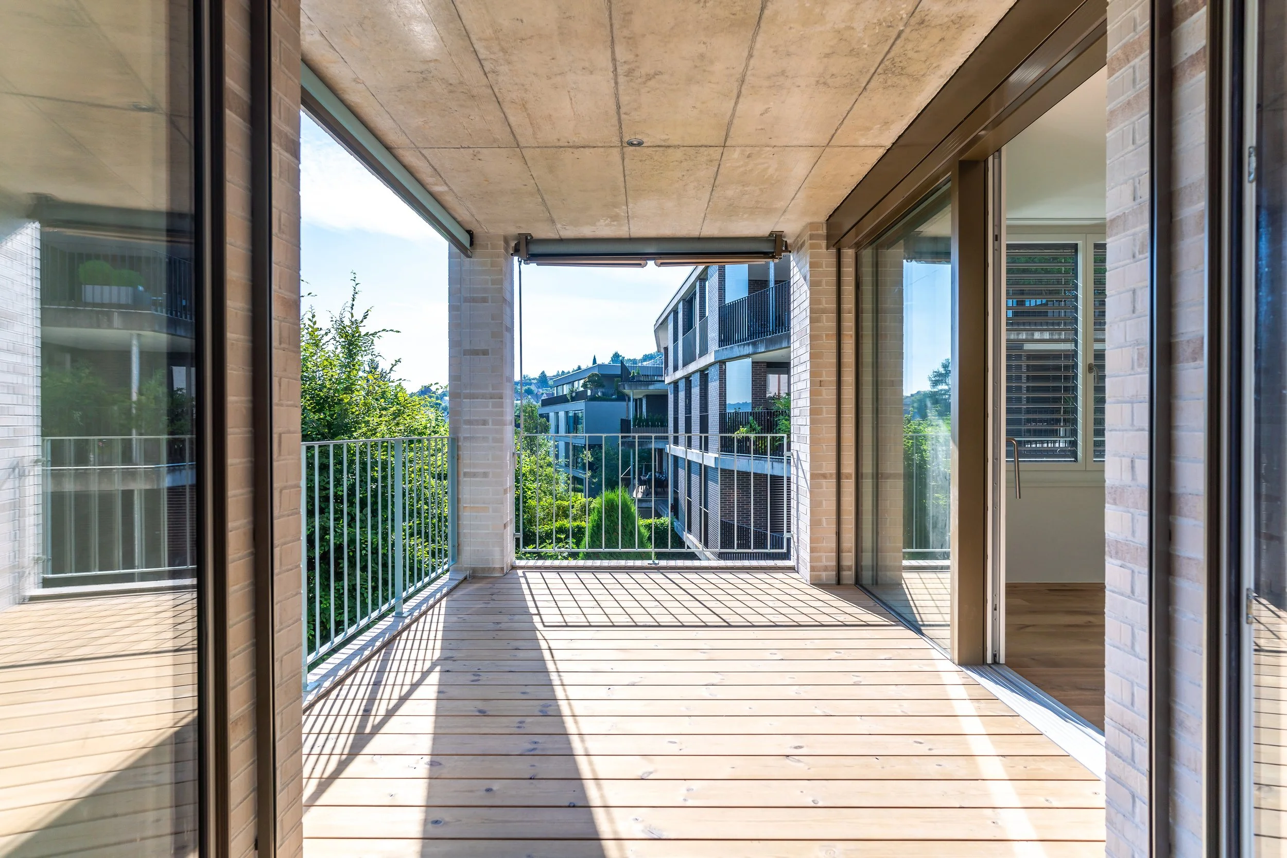 Balkon mit Holzdecke, Flügeltüren und Blick auf moderne Nachbargebäude, umgeben von Bäumen.