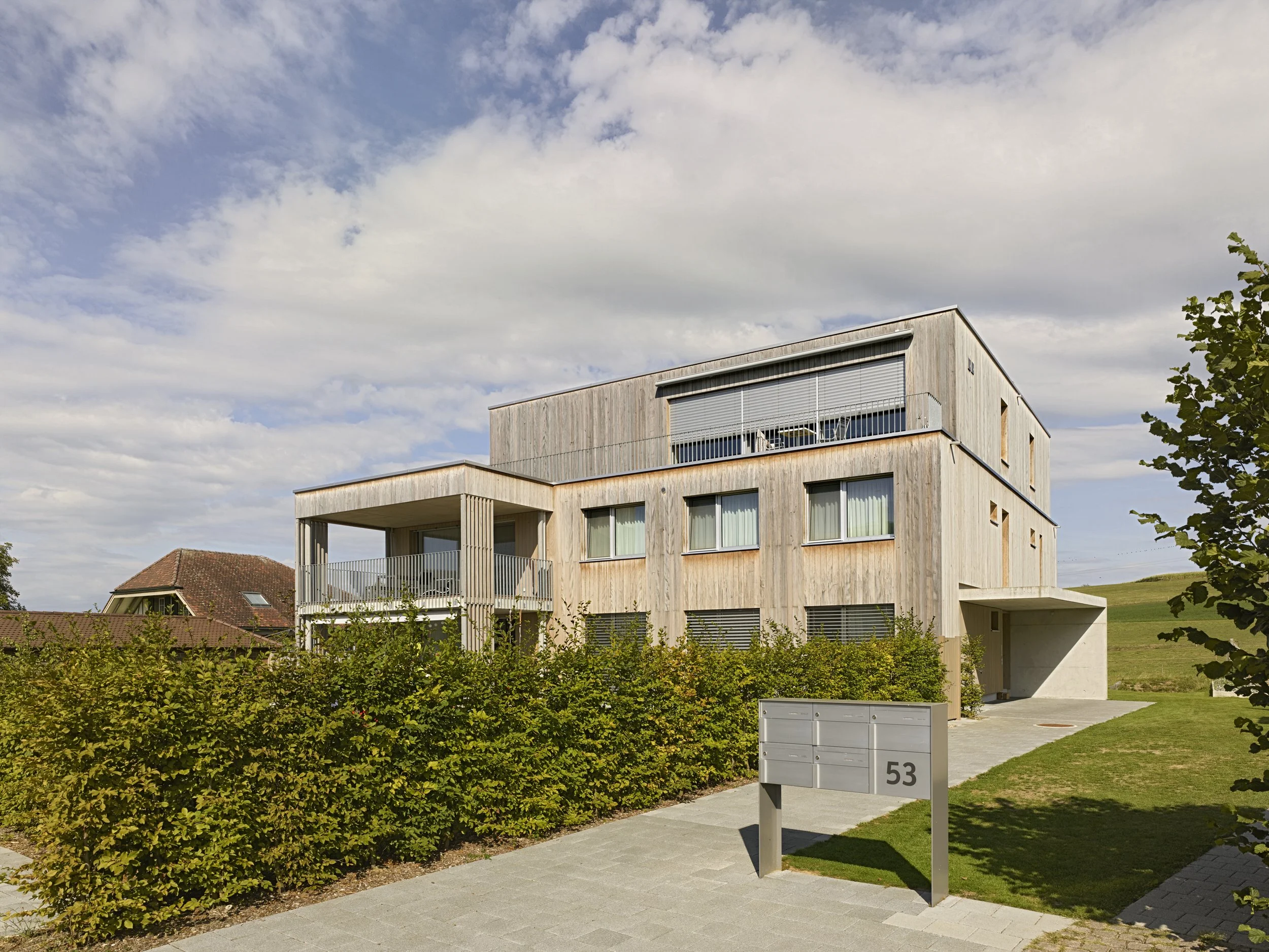 Modernes, mehrstöckiges Wohngebäude mit Holzfassade und großen Fenstern, umgeben von Grünflächen und einem Gehweg; im Vordergrund ein Baum und eine Hausnummer 53