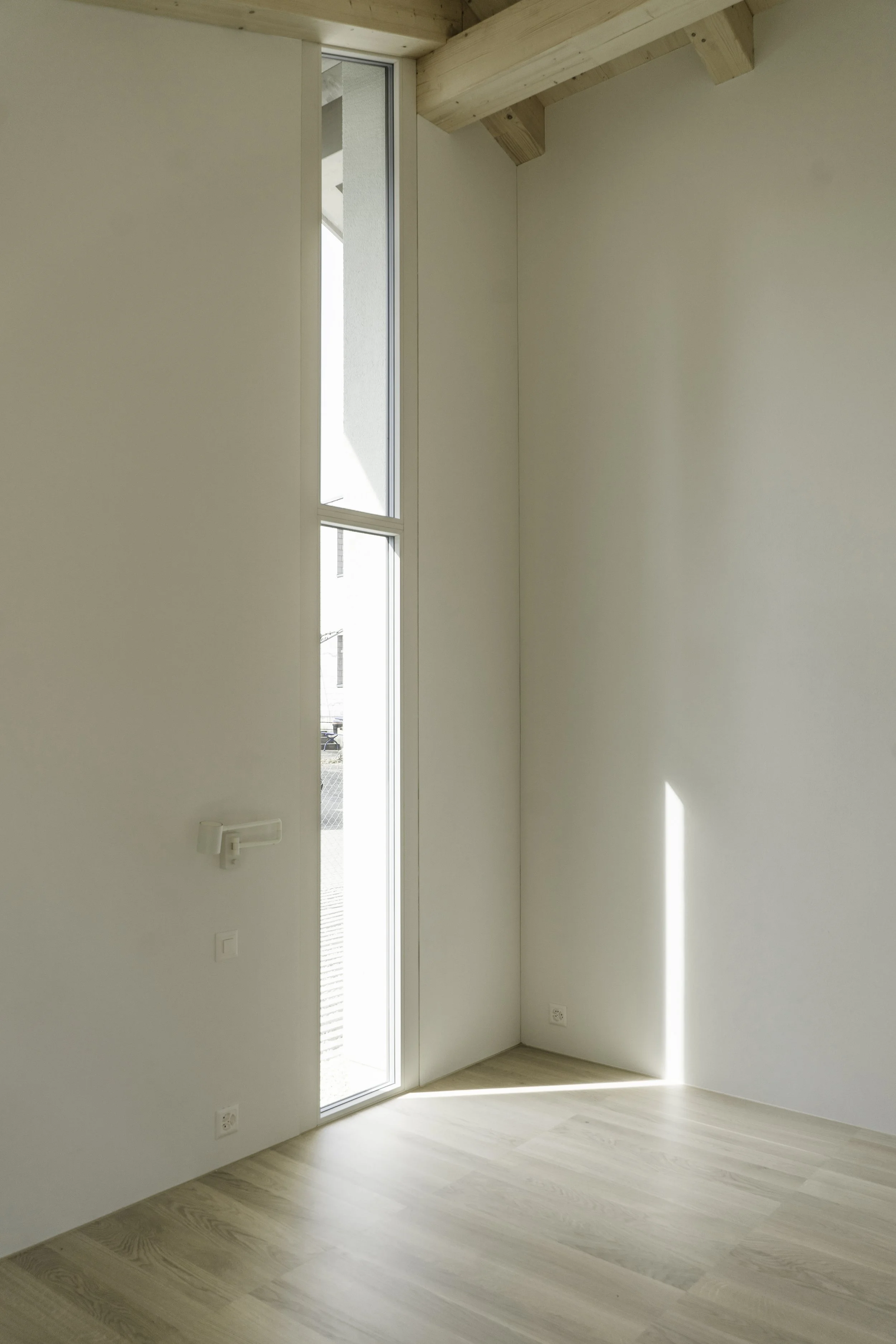 Ein heller, leerer Raum mit weißen Wänden, Holzdielenboden und einem hohen Fenster, das Licht hereinlässt. Der Raum hat eine Dachschräge mit sichtbaren Holzbalken.