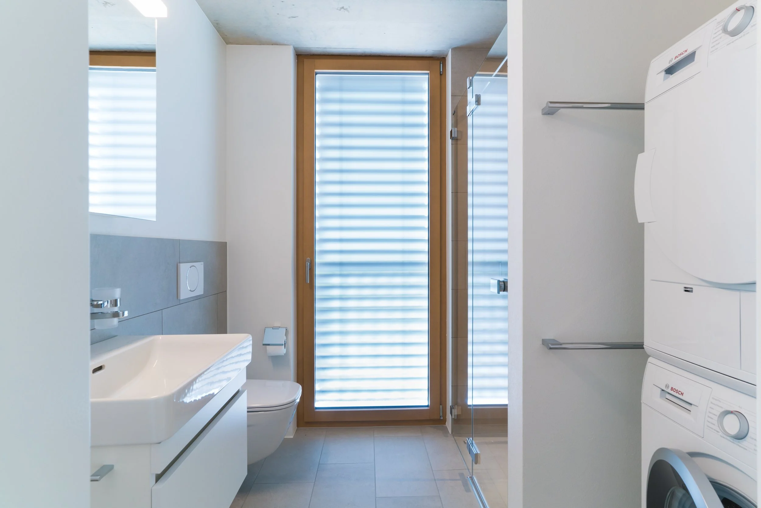 Ein moderner Waschrraum mit Waschbecken, Toilette, Waschmaschine und Trockner, sowie eine große Fenstertür aus Holz.