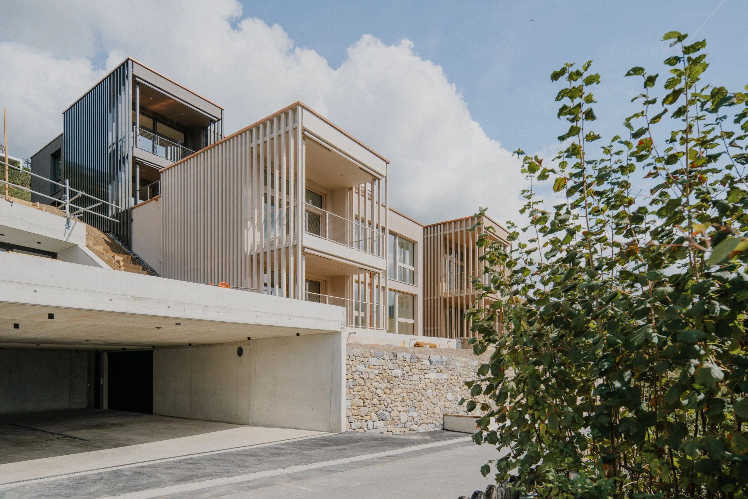 Moderne mehrstöckige Wohnhäuser mit Balkonen und Holzverkleidungen, angrenzend an einen Parkplatz und einen Garten mit grünen Bäumen, bei sonnigem Himmel.