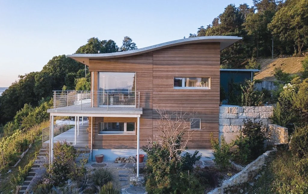 Moderner Holzhauss mit Balkon und terrassiertem Garten, umgeben von Bäumen und Natur, auf hügeliger Landschaft gelegen.