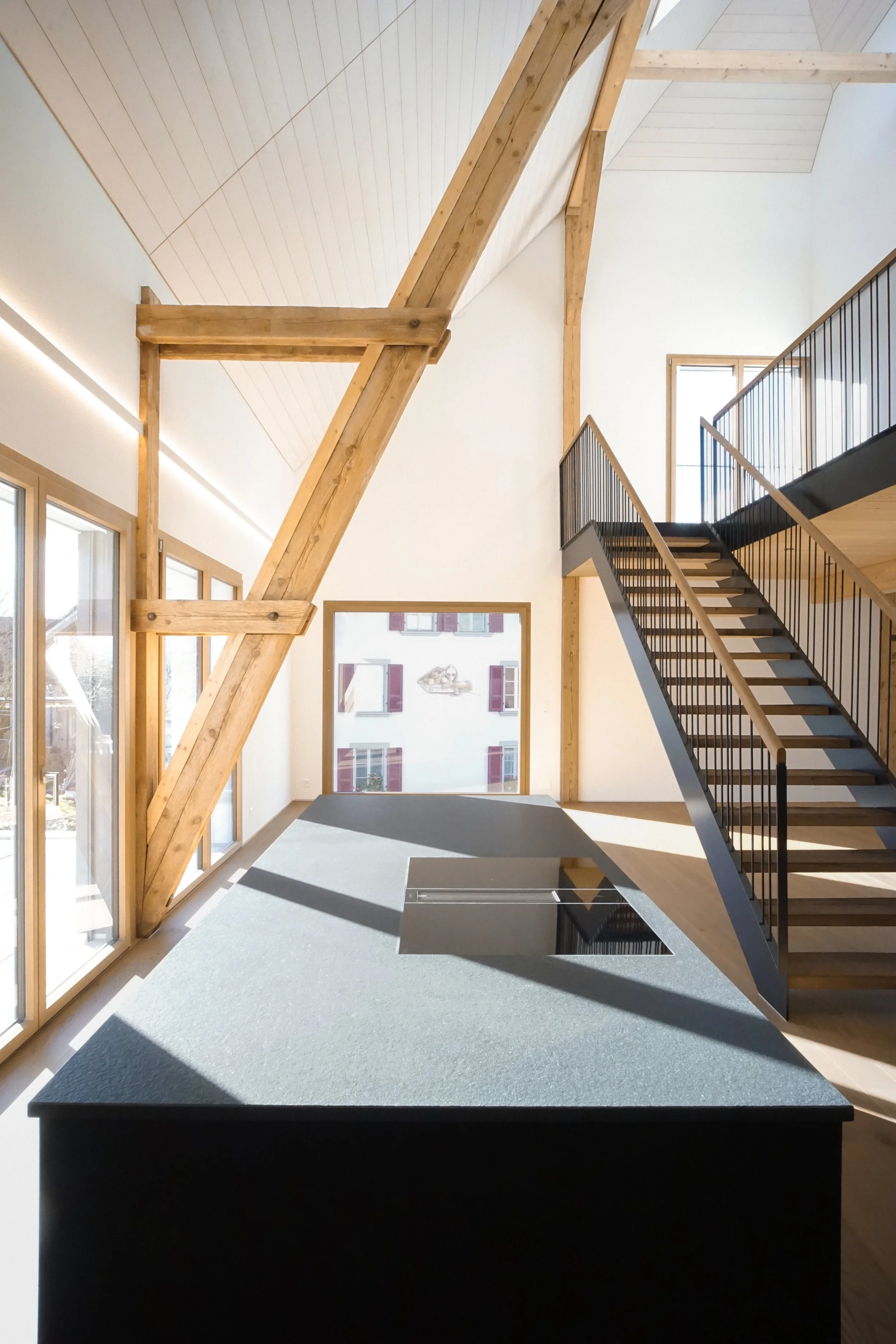Ein heller, moderner Raum mit großen Fenstern, Holztragwerken, einer Treppe mit schwarzem Geländer und einem schwarzen Tisch mit Loch in der Mitte.