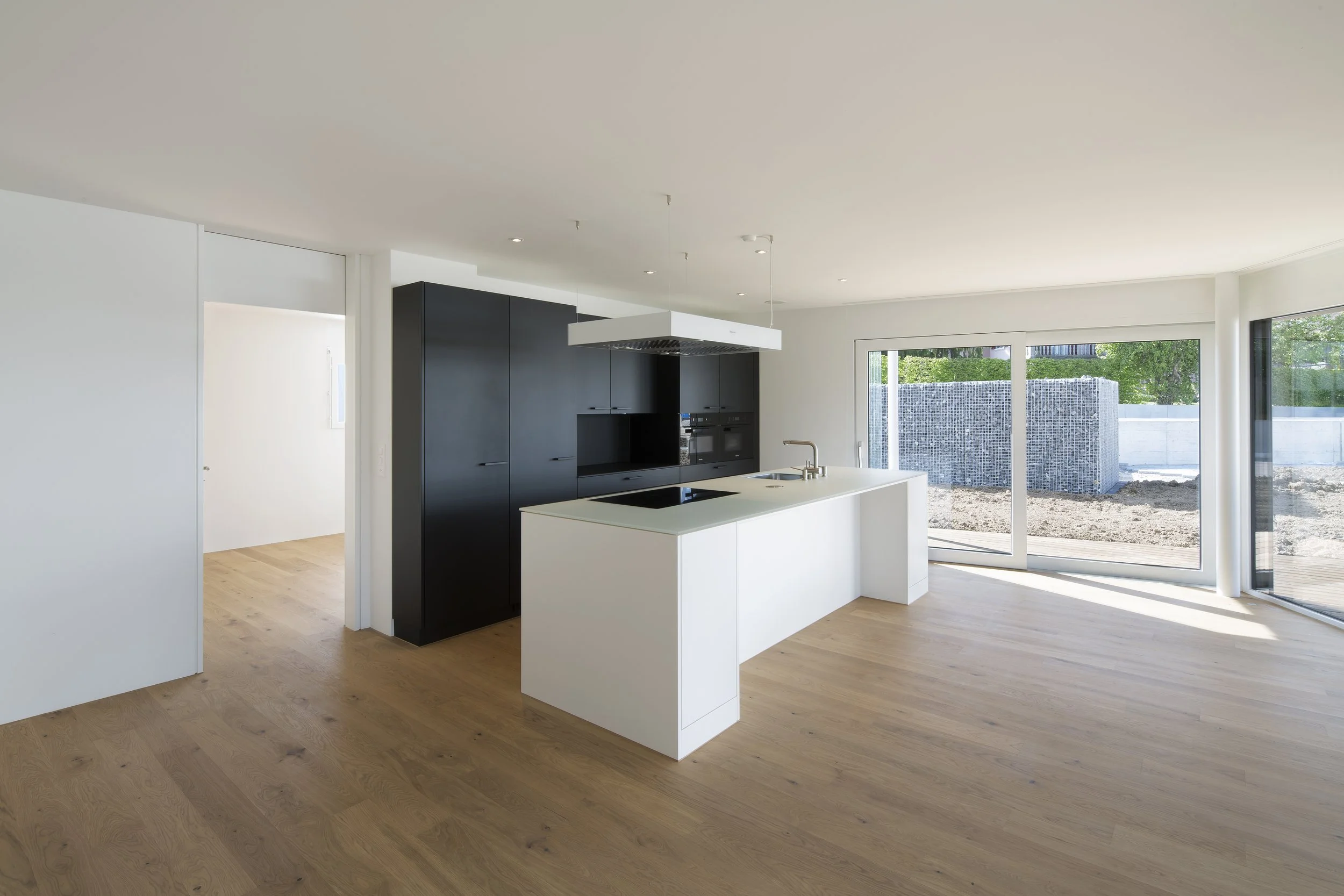 Moderne offene Küche mit weißer Insel, schwarze Einbauküche, große Fenster mit Blick nach draußen, helle Holzboden.