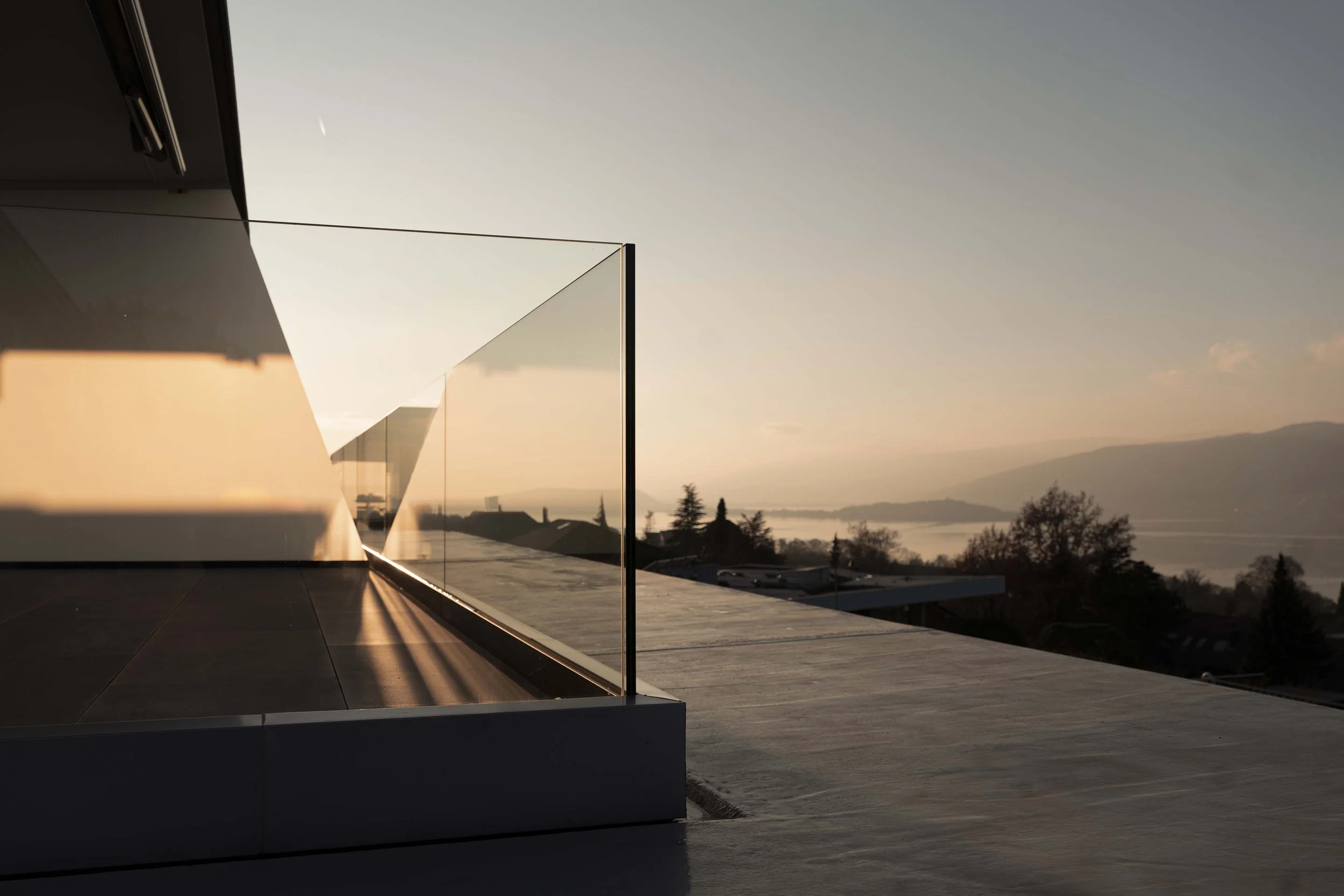 Moderner Balkon mit Glasgeländer bei Sonnenuntergang, Blick auf Berge und Wasser im Hintergrund.