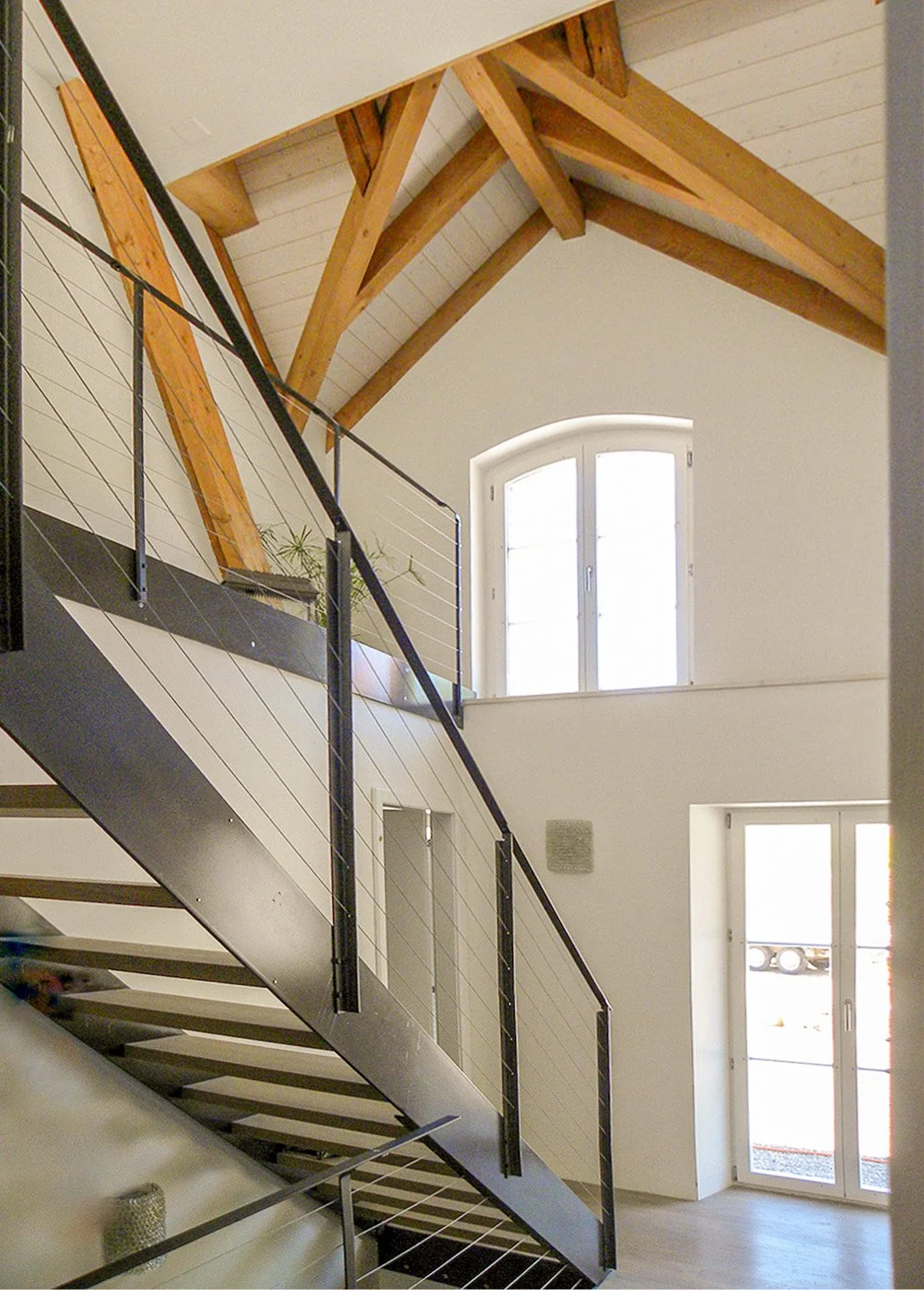 Innenansicht eines Hauses mit einer modernen Treppe, großen Fenstern, weißen Wänden und sichtbaren Holz-Dachbalken.