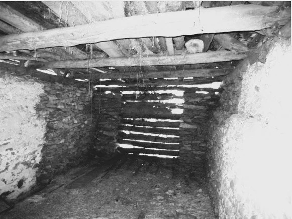 Innenansicht eines alten, ruinenartigen Steinschuppens mit Holzdach und schmalem Eingang, Licht fällt durch die Ritzen zwischen den Steinen.