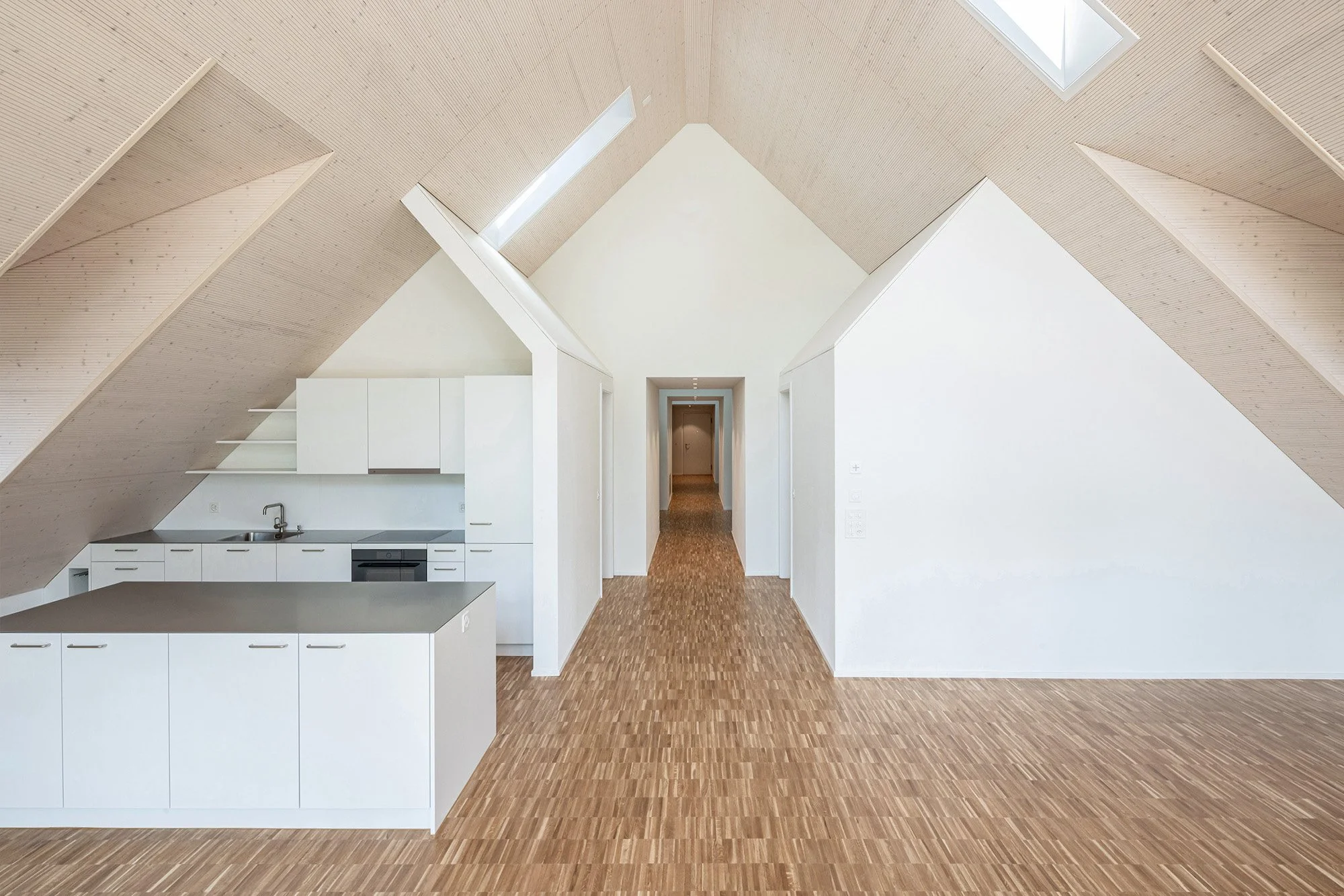 Helles, minimalistisches Zimmer mit weißen Wänden, einer Küchenzeile und einem hohen, schrägen Decke mit Dachfenstern, Holzboden.