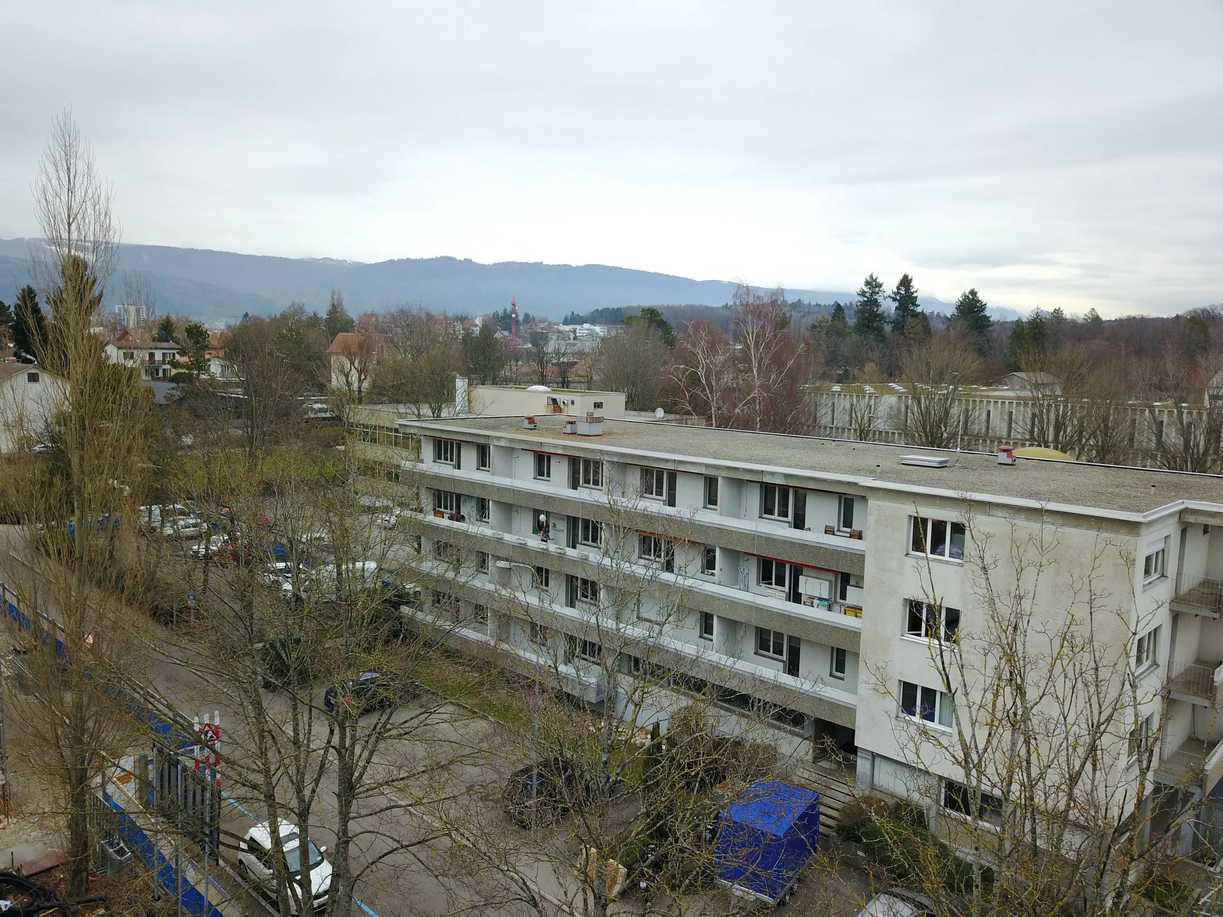 Blick auf eine Wohnanlage mit mehreren weißen, mehrstöckigen Gebäuden und geparkten Autos im Vordergrund. Im Hintergrund sind Bäume, Berge und ein bewölkter Himmel sichtbar.