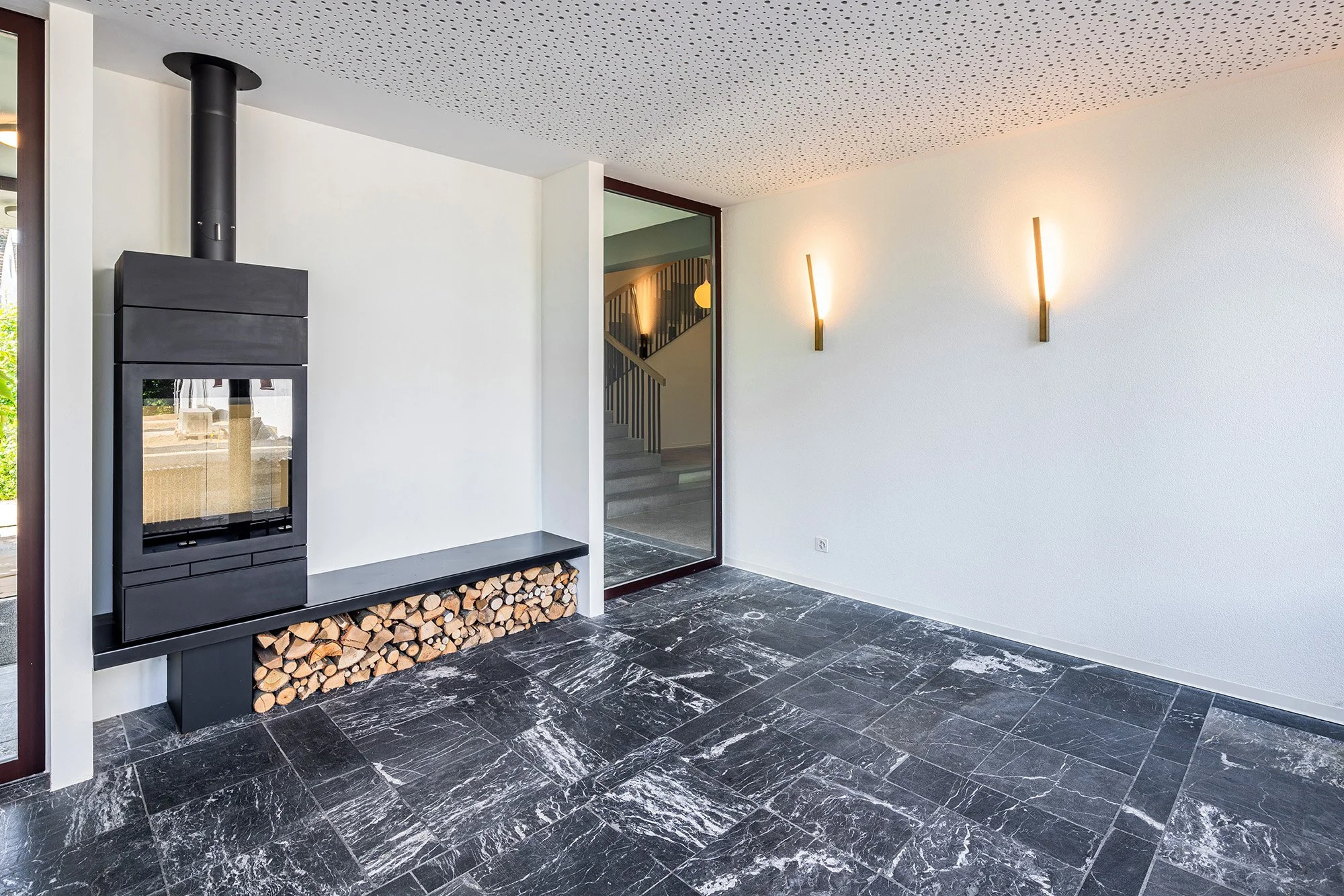 Innenraum einer modernen Wohnung mit schwarzem Holzofen, Feuerholz unter einer Ablage, weißen Wänden, dunklem Marmorfußboden, und Wandlampen.