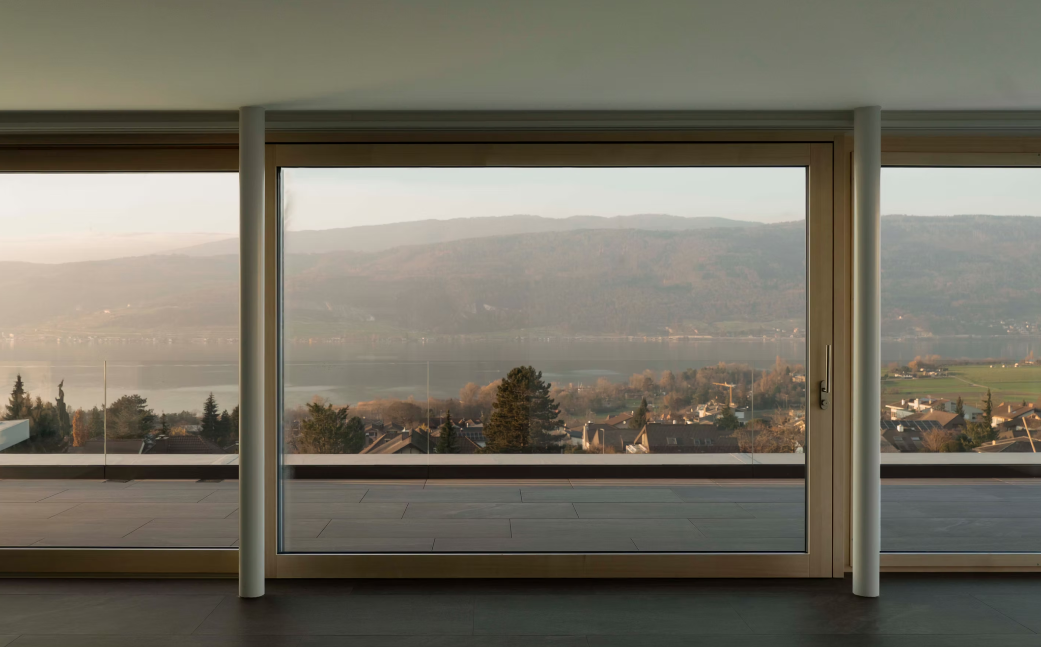 Blick durch eine große Fensterfront auf einen See, bewaldete Hügel und ein Dorf. Innen sind weiße Säulen und ein dunkler Boden sichtbar.