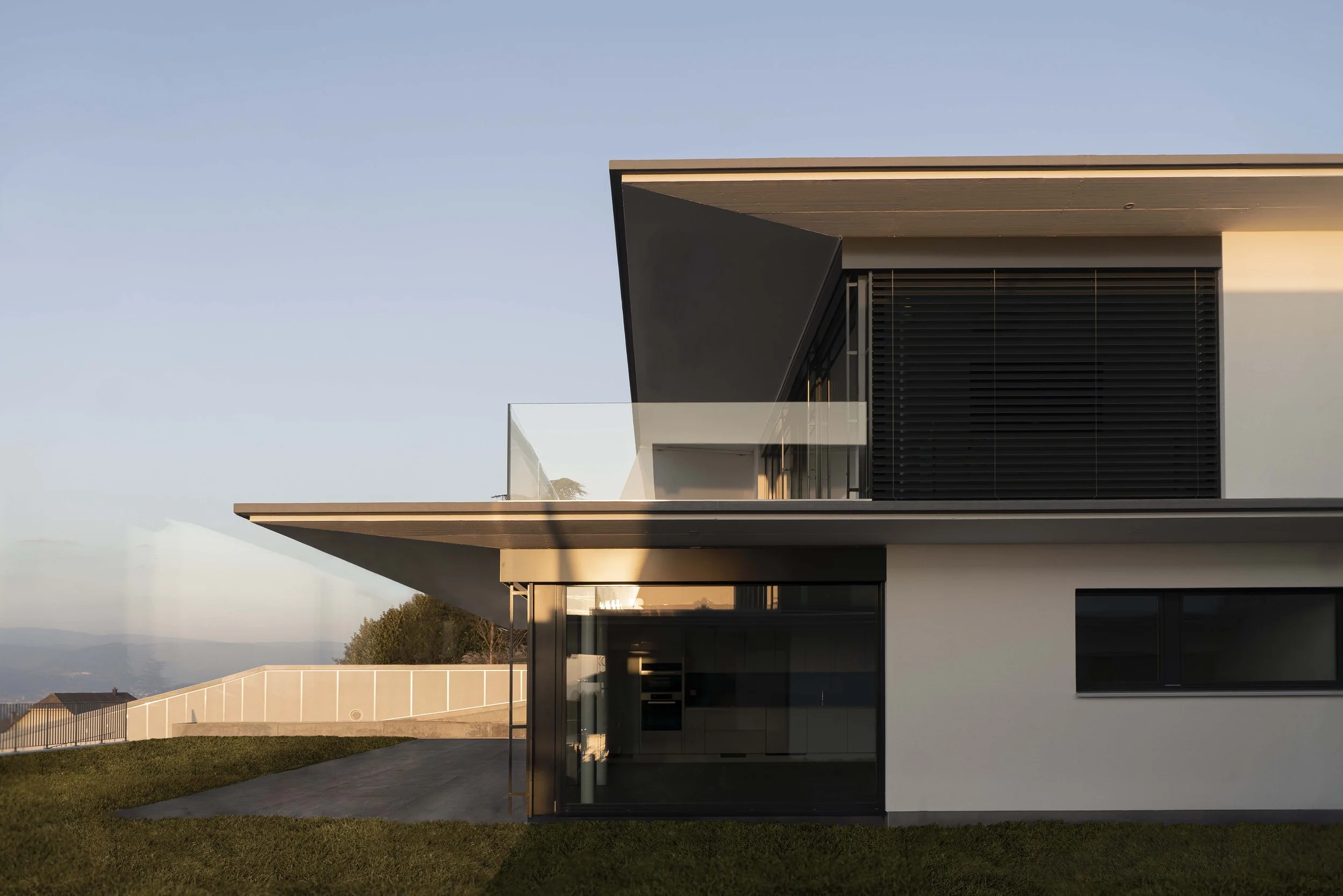 Modernes, mehrstöckiges Haus mit großen Fenstern, einer Terrasse mit Glasgeländer und einem minimalistischen Design, umgeben von einem gepflegten Rasen, bei Sonnenuntergang.