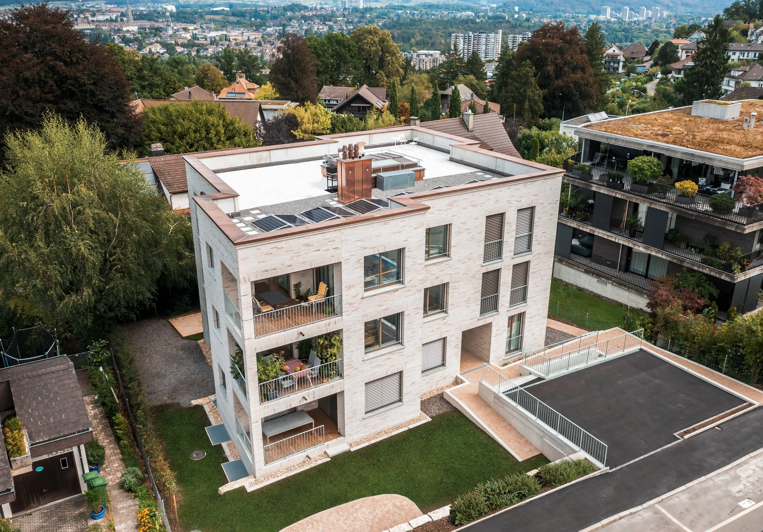 Ein moderner, weißer Wohnkomplex mit mehreren Balkonen und Solarzellen auf dem Dach, umgeben von Bäumen und einer städtischen Umgebung.