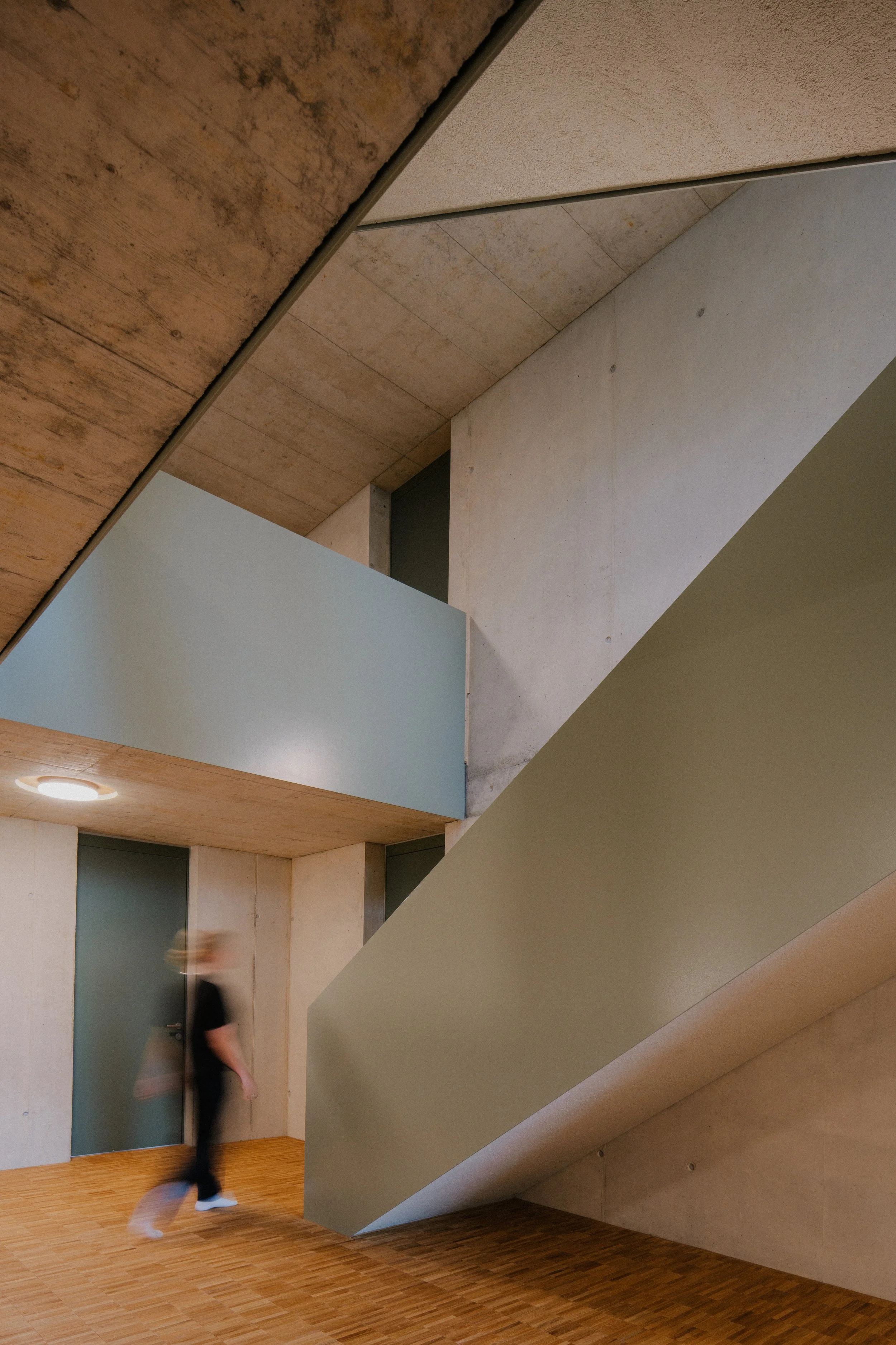 Innenaufnahme eines Gebäudes mit modernen, geometrischen Architekturelementen, Sicht auf eine Treppe und eine Person, die geht.