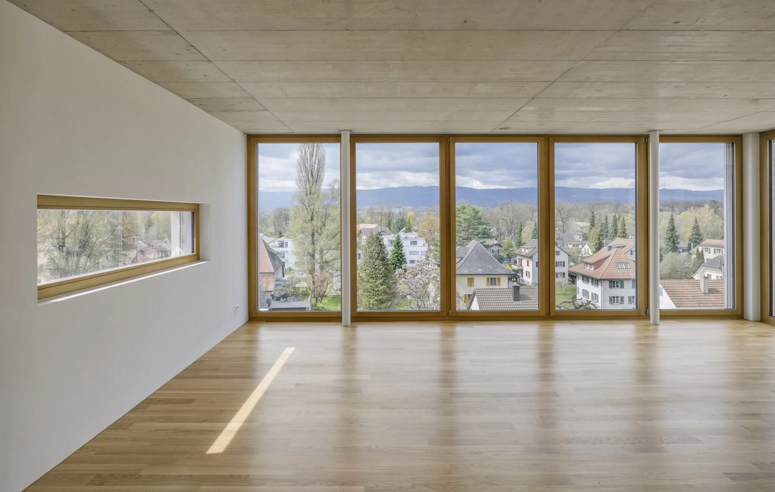 Innenraum eines modernen, leeren Zimmers mit großen Fensterfronten und Holzboden, Blick auf eine Stadtsiedlung mit Häusern und Bäumen unter einem bewölkten Himmel.