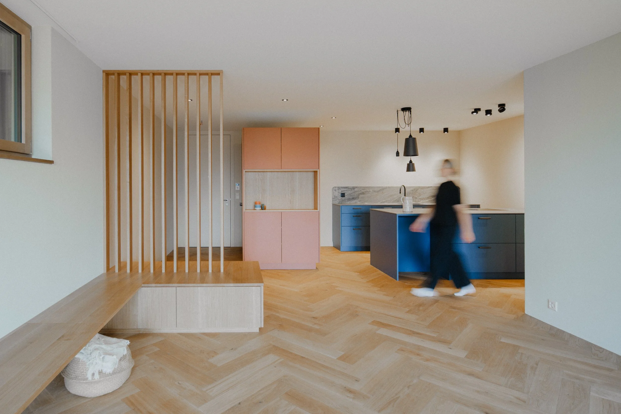Moderner Küchenraum mit blauer Kücheninsel, rosa Schränken, Holzboden und Lichtelementen, mit einer Person in Bewegung.