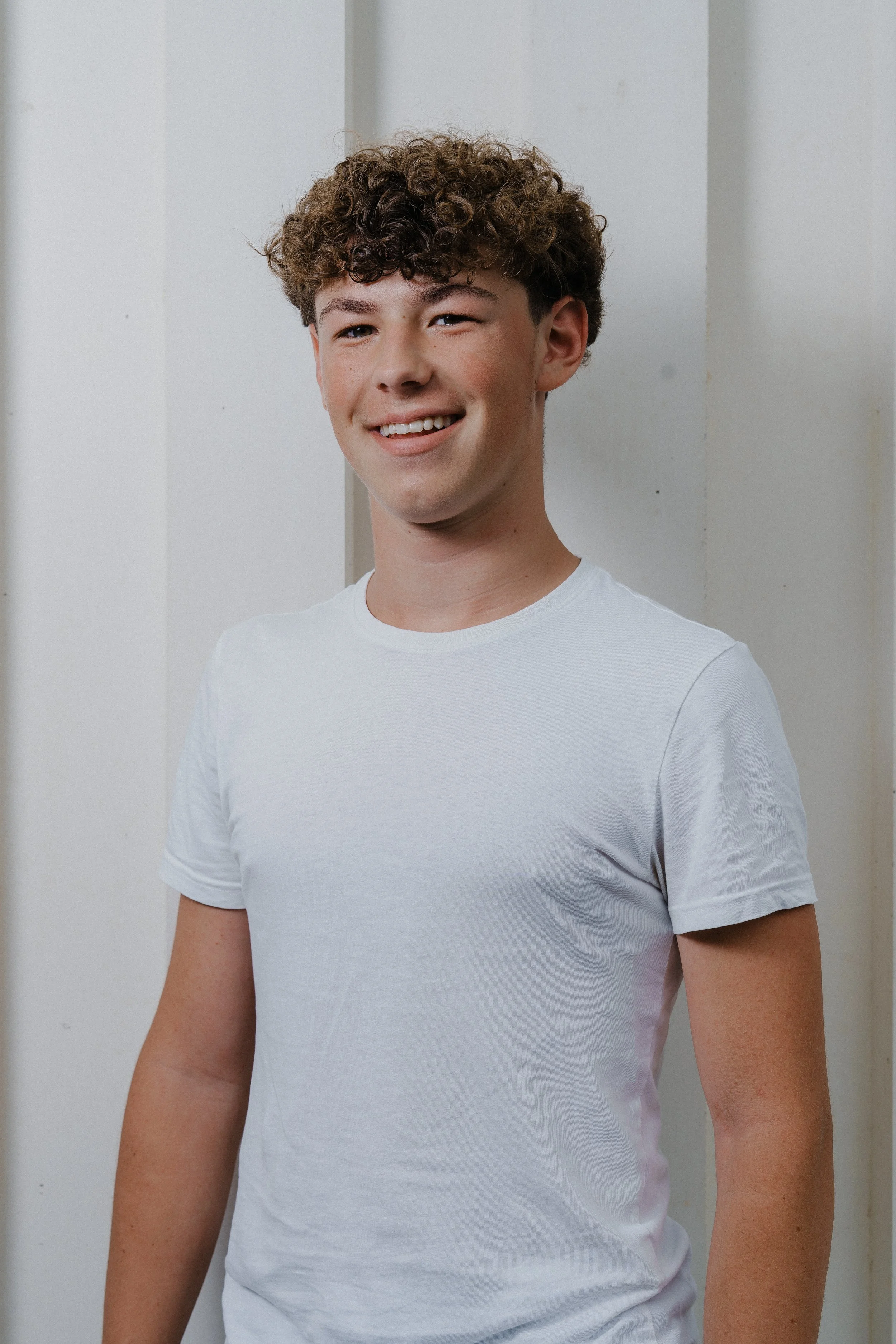 Junge mit lockigem braunem Haar, lächelnd, in einem weißen T-Shirt stehend vor einer weißen Wand.