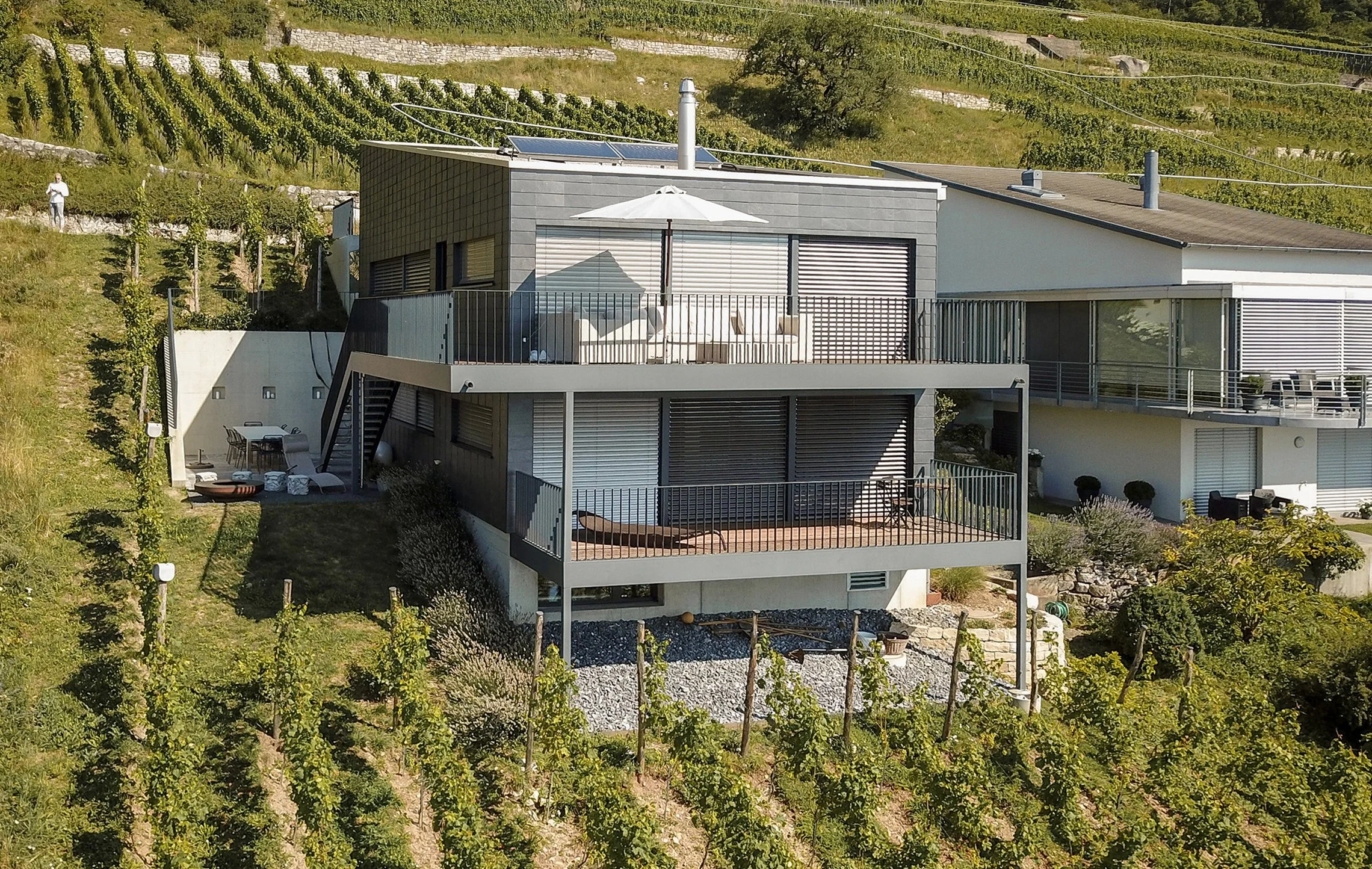 Moderne mehrstöckige Villa auf einem Hügel, umgeben von Weinbergen und Garten, mit Terrasse und Sonnenliege im Garten, bei sonnigem Wetter.