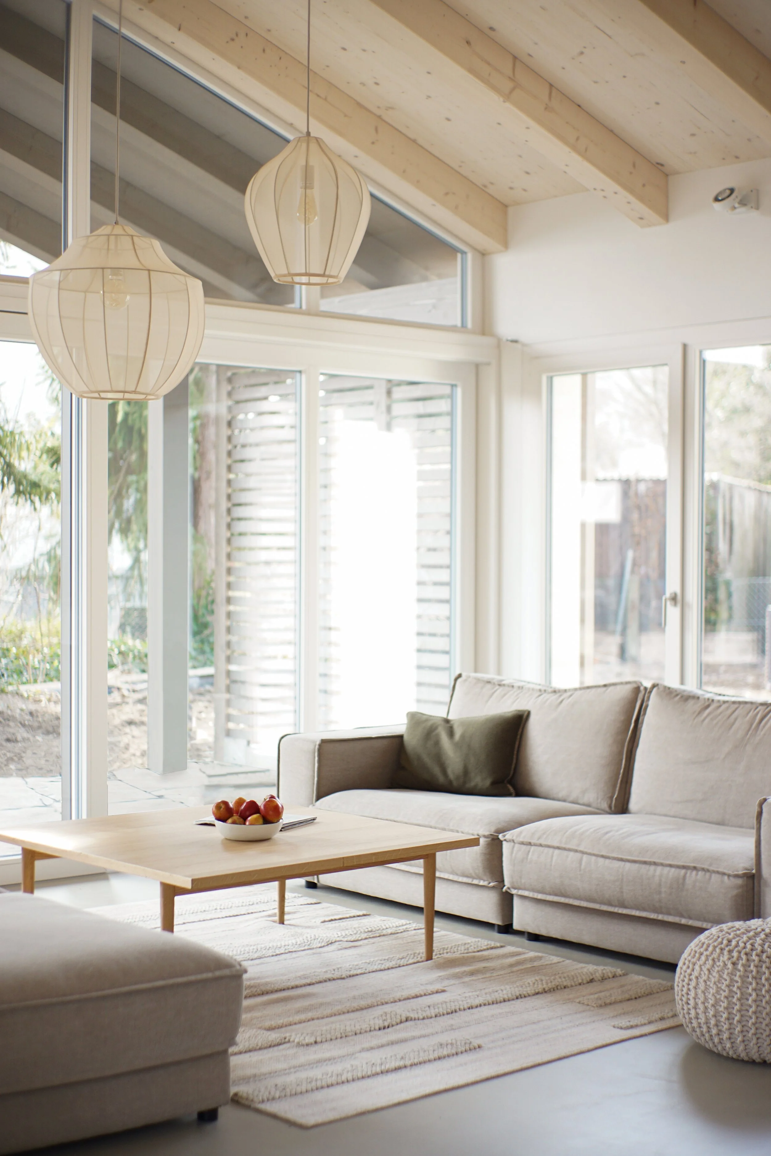 Heller Wohnraum mit großen Fenstern, weißem Sofa, Kaffeetisch mit Obst, Decken, Holzdecke, Hängelampen, Gardinen und Blick nach draußen.