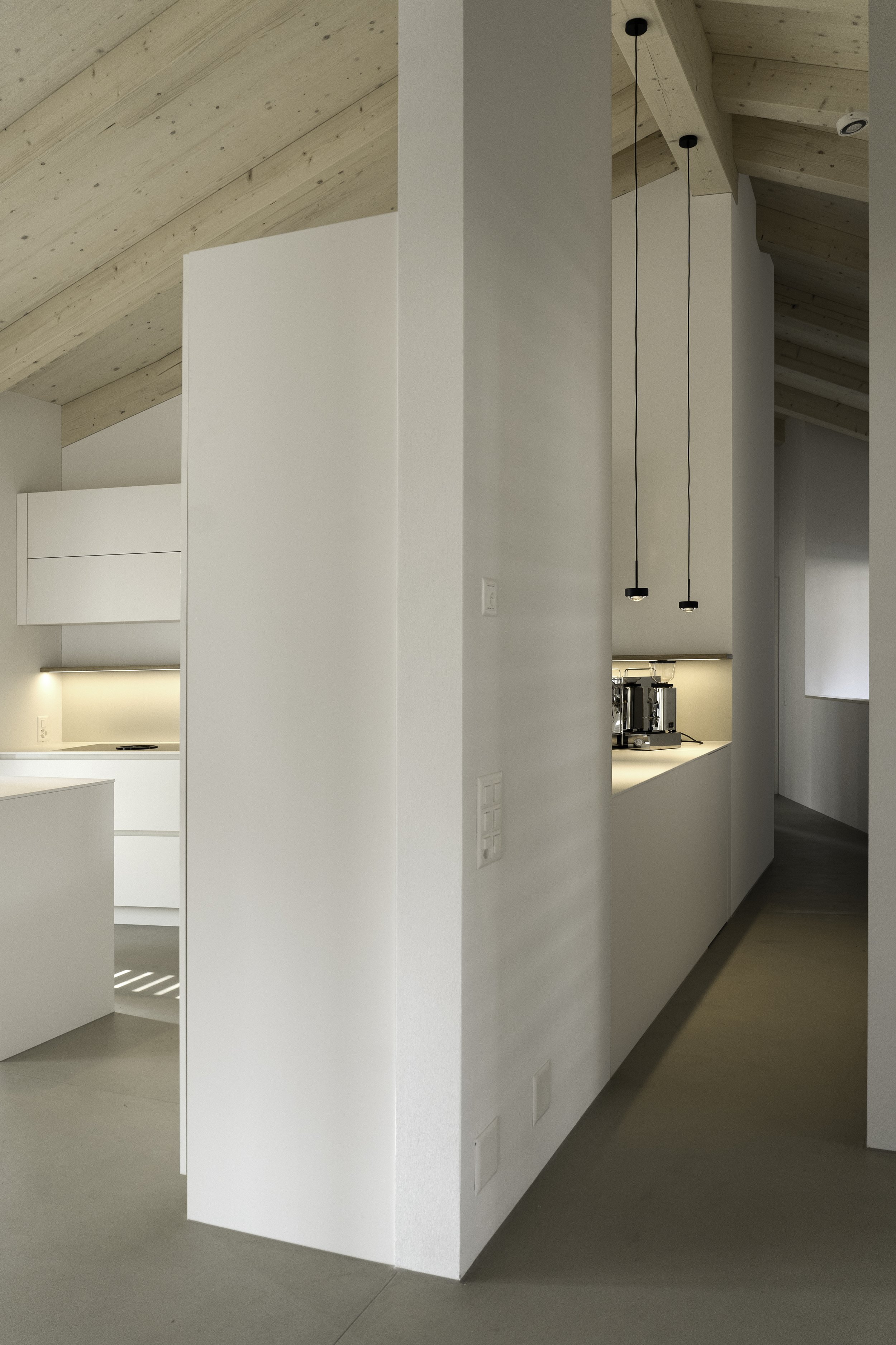 Innenraum eines modernen Hauses mit weißen Wänden, Holzdecke und minimalistischem Design, inklusive Pendelleuchten und Kochausschnitt im Hintergrund.
