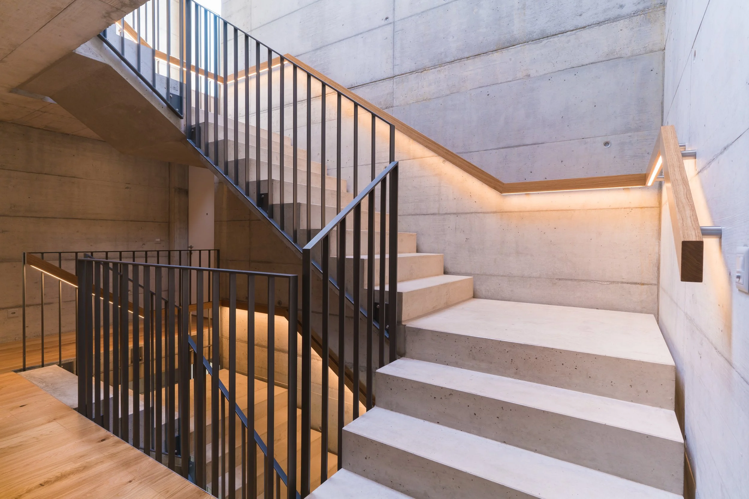 Innenaufnahme einer modernen, mehrstöckigen Treppe mit schwarzem Metallgeländer und Holzhandlauf in einem Gebäude mit rohem Betonwänden und -boden, gedämpfter Beleuchtung entlang der Treppenkanten.