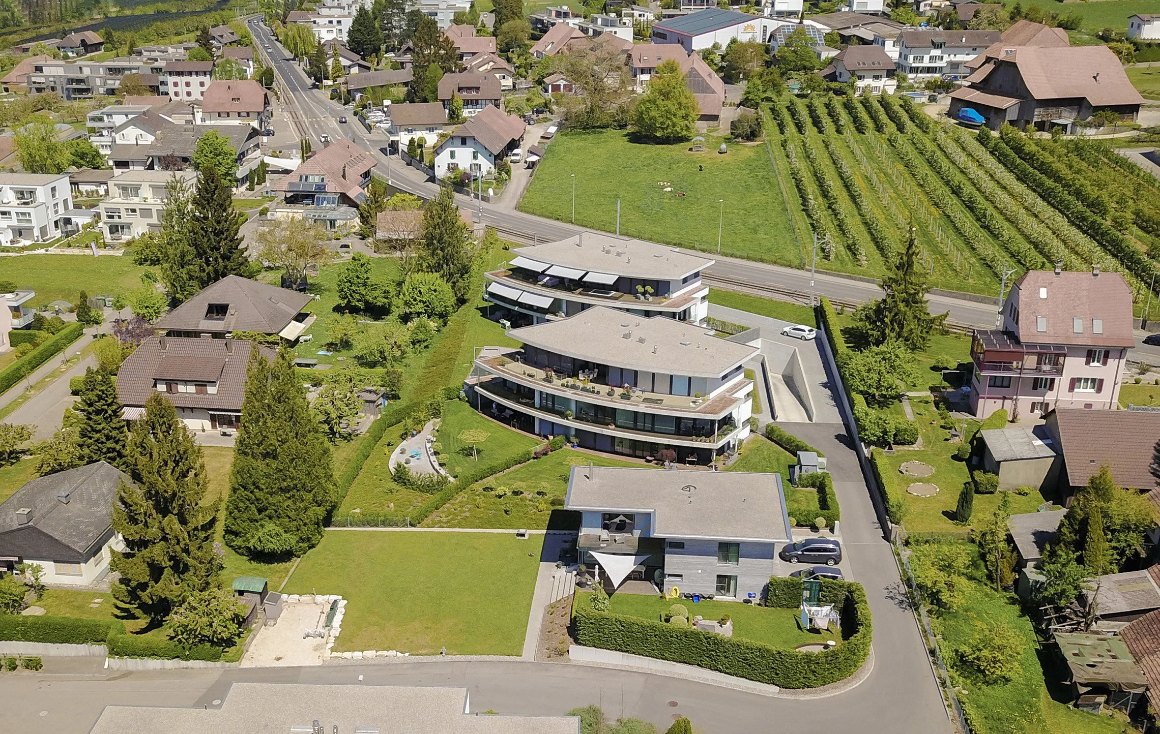 Luftaufnahme eines Wohngebietes mit mehreren Häusern, Gärten und einem modernen Mehrfamilienhaus, umgeben von Bäumen und Wiesen.