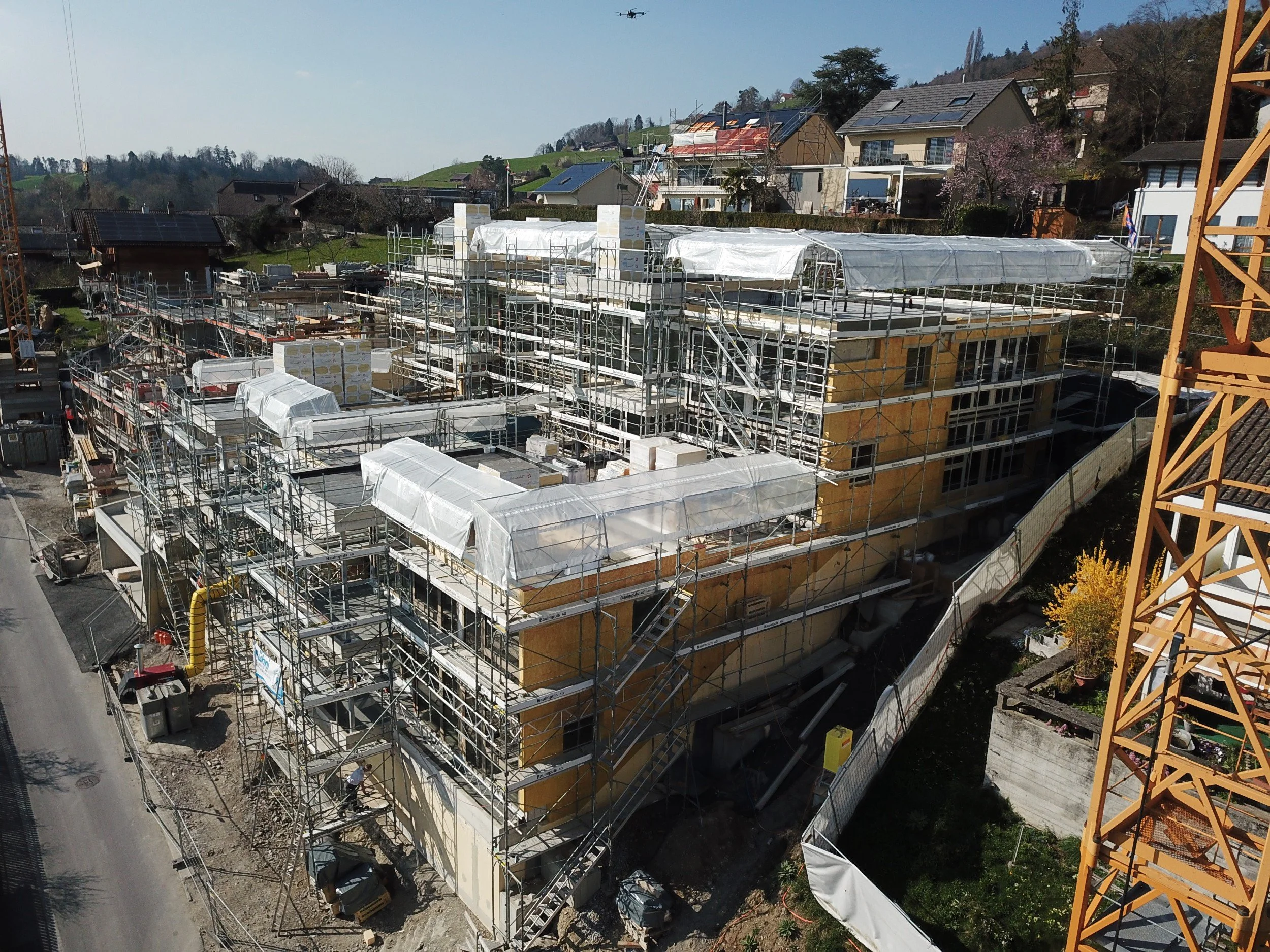 Baustelle in einem Wohngebiet mit mehreren eingeschlagenen Gebäuden, Baugerüsten, Bauhelfern und einem Kran. Im Hintergrund sind Häuser mit Solardächern und eine grüne Hügellandschaft.