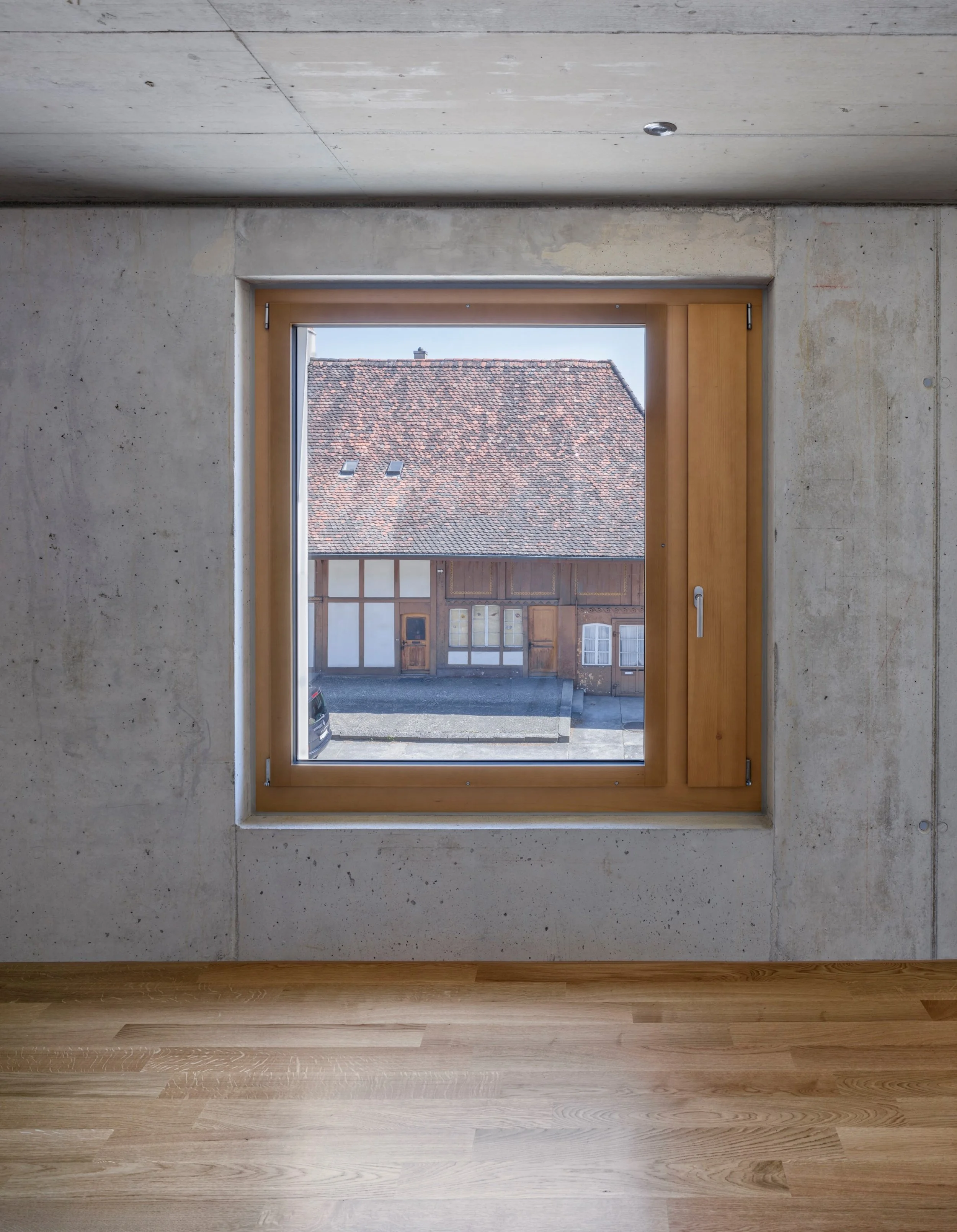 Innenaufnahme eines Zimmers mit einem großen Fenster, das auf ein Dach mit roten Ziegeln und zwei kleine Gauben blickt. Die Wand um das Fenster ist aus unbehandeltem Beton, der Boden aus hellem Holz.