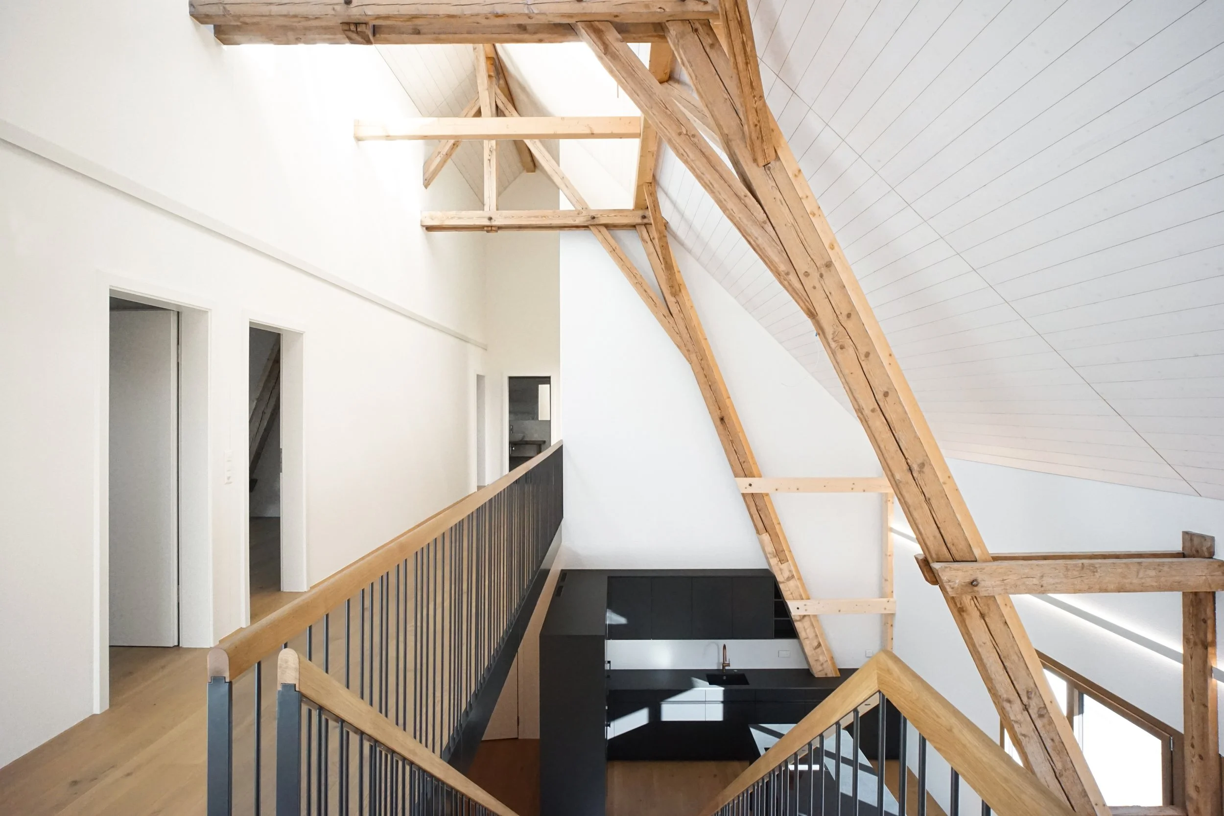 Innenaufnahme eines modernen Hauses mit sichtbaren Holzdecken und offener Treppe mit Holzhandlauf und schwarzen Balustraden, weiße Wände und mehrere Türen.