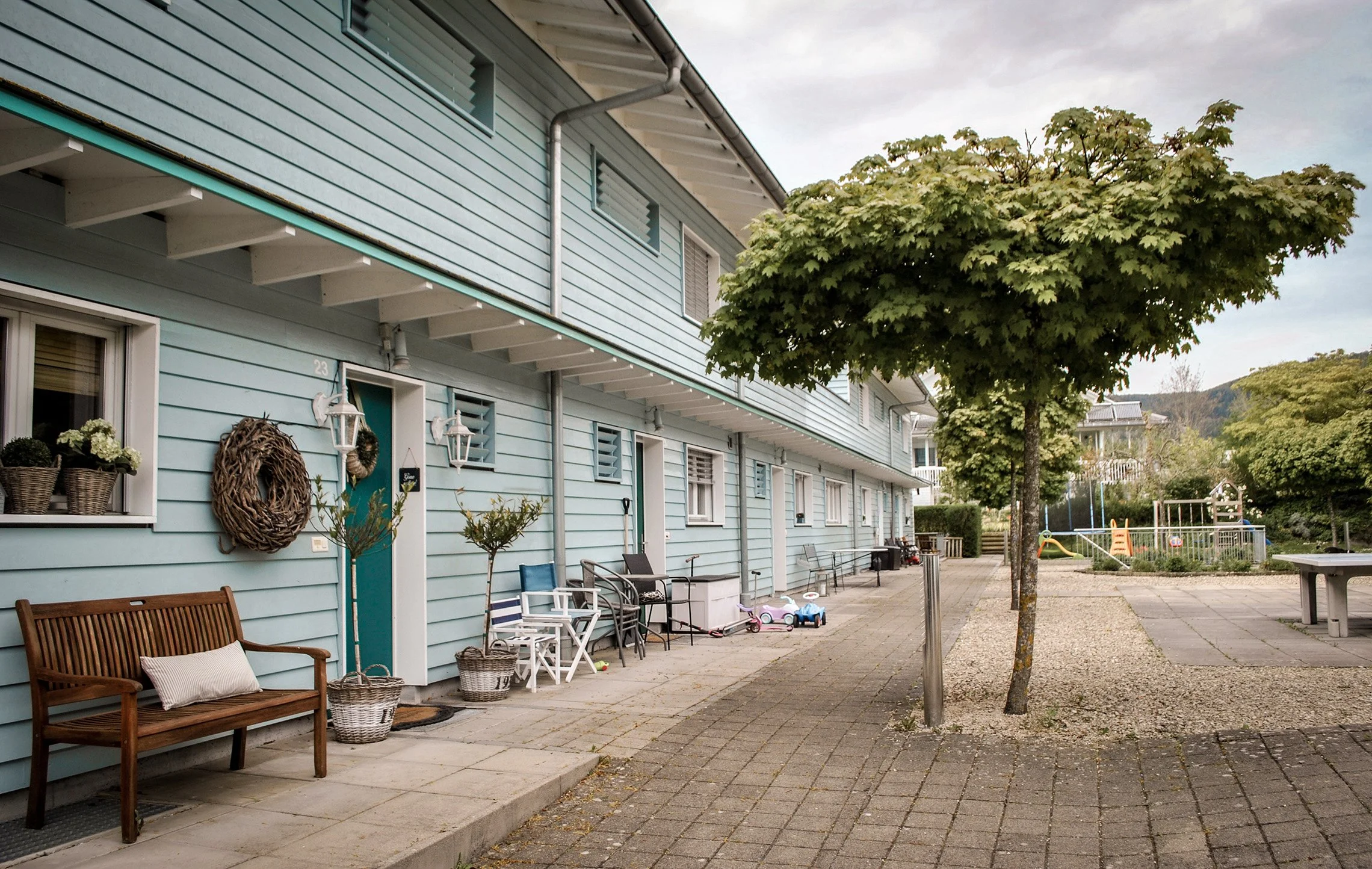 Ein blauer Mehrfamilienhaus mit vorgarten und Spielplatz im vordergrund, Bäume und ein bewölkter Himmel