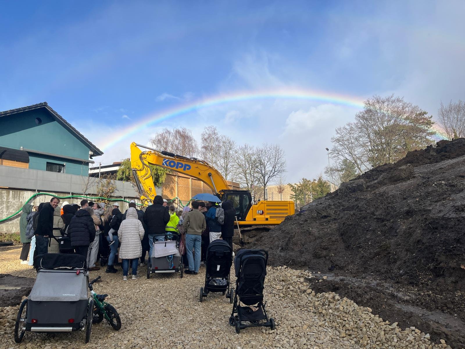 Gruppe von Menschen auf einer Baustelle mit Bagger, hunderten von Reisetaschen und Kinderwagen, Beobachtung des Bauvorhabens bei Regen unter Regenbogen, draußen in einer urbanen Umgebung mit Häusern und Bäumen.